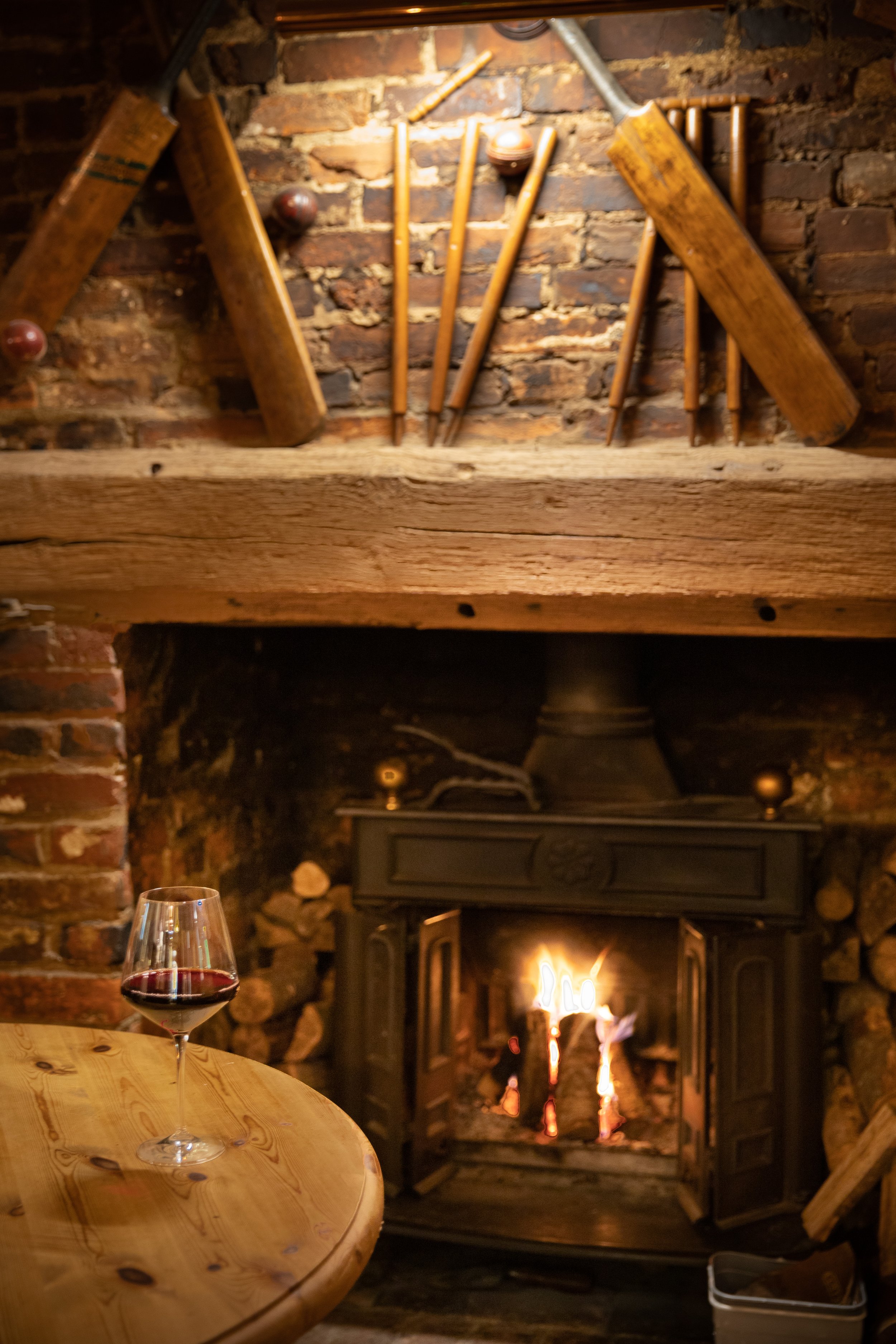 A cozy fireplace with burning logs, a wooden table with a glass of red wine, and vintage woodworking tools hanging on a brick wall.
