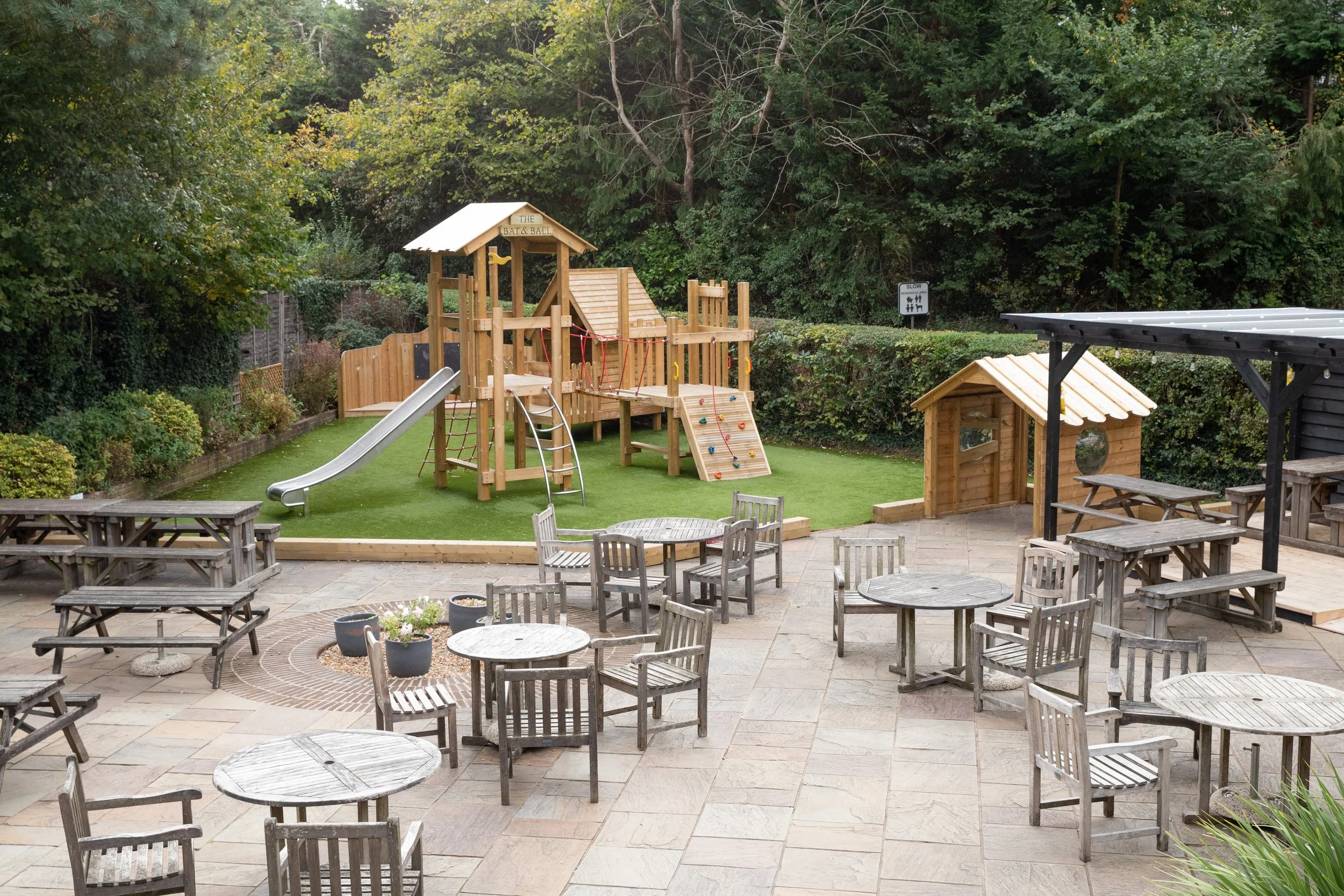 An outdoor children's playground with a wooden play structure, slide, climbing wall, and rope bridges, surrounded by a grassy area with trees and bushes. There are several wooden tables and chairs on a paved patio area next to the playground.