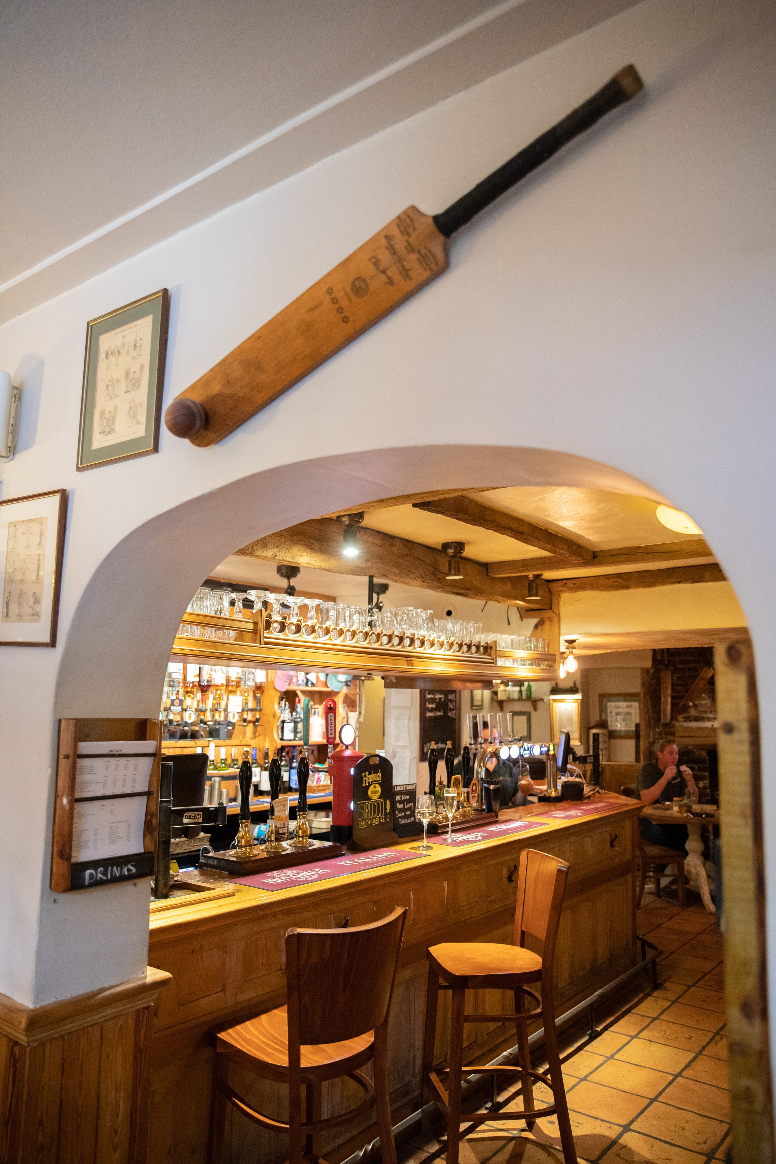 A pub with a bar counter made of wood, beer taps, and glasses hanging above. There are two wooden chairs, and a person sitting at a table in the background.