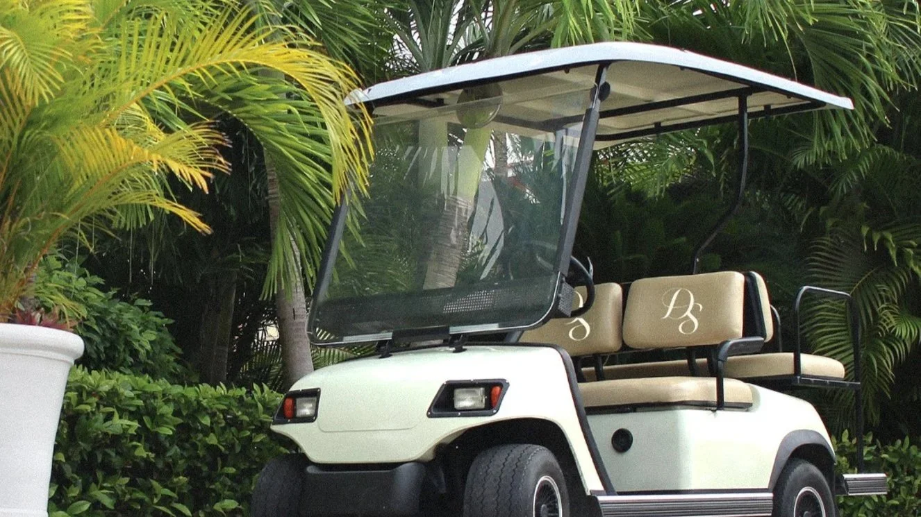 A white luxury golf cart parked in a tropical garden setting with palm trees, representing the private resort transportation service