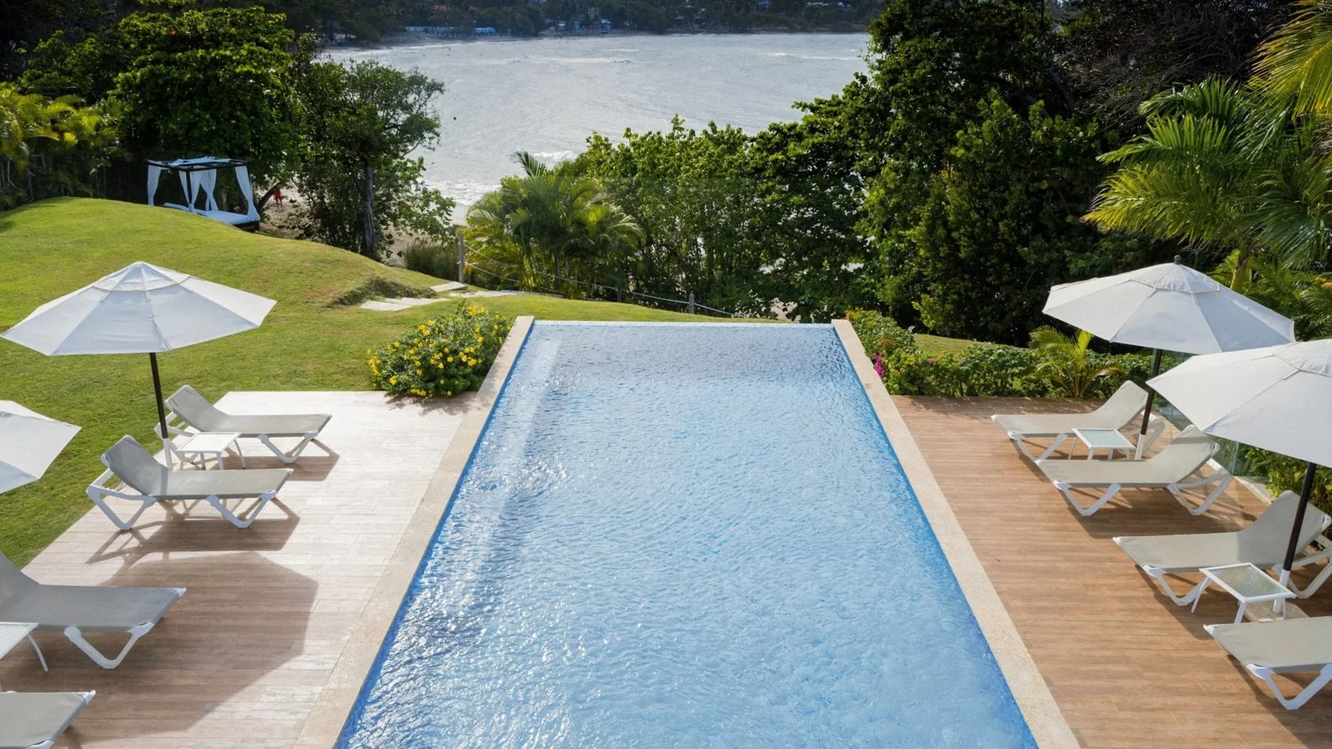 Infinity pool with white sunbeds and umbrellas, surrounded by lawn and greenery, overlooking a river or lake in the background.