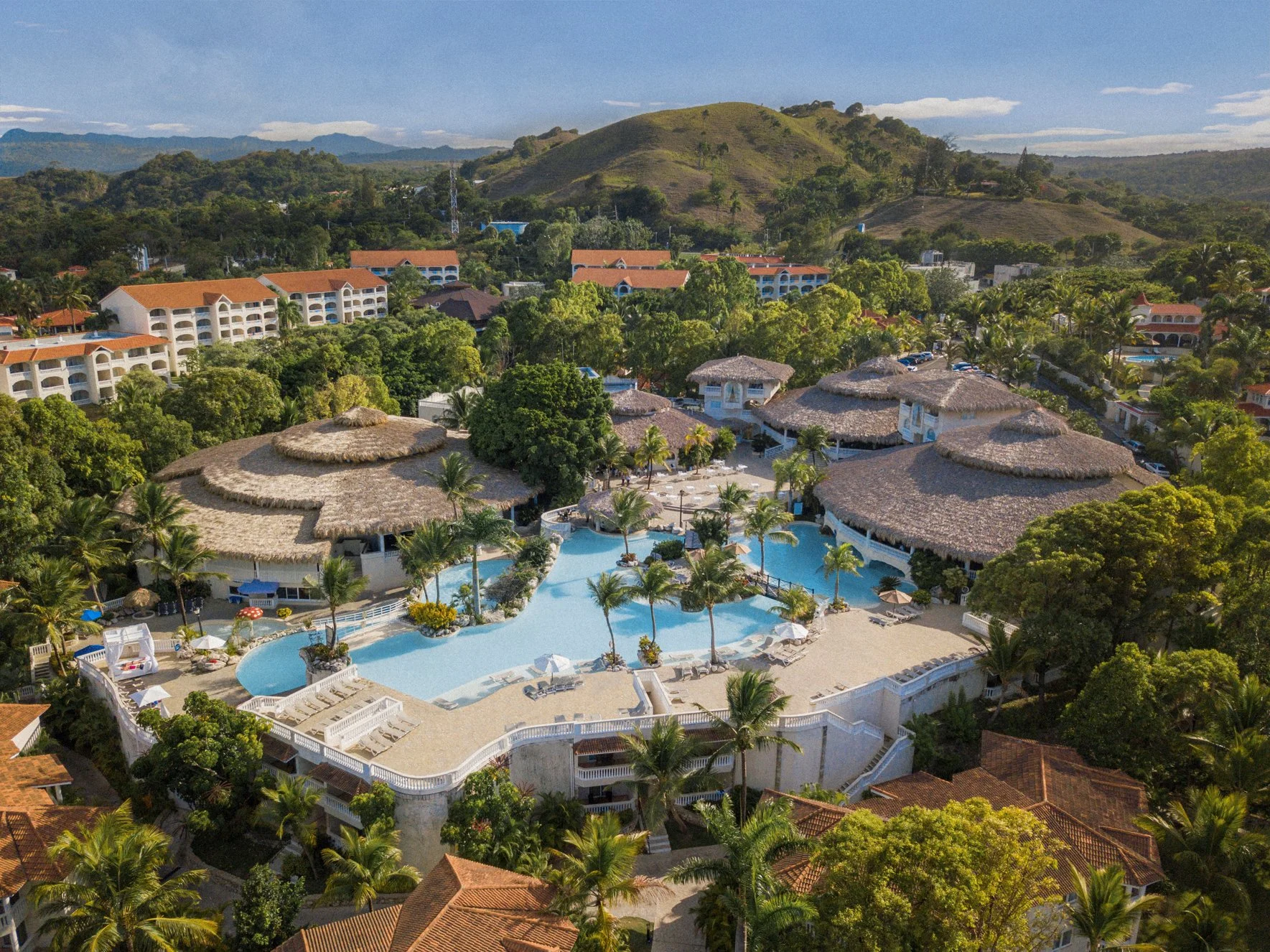 Aerial view of a luxury Caribbean resort featuring sprawling turquoise pools, tropical palm trees, and elegant villas with thatched roofs under a clear blue sky.