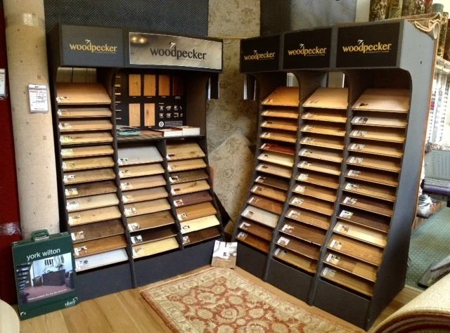 Display of wood samples in a retail store with Woodpecker branding, arranged in slanted racks with various shades of wood finish.