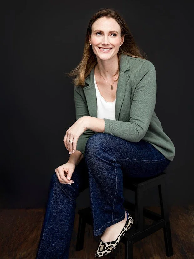 A woman with long brown hair, smiling, sitting on a black stool against a dark background. She is wearing a green blazer, white top, blue jeans, and leopard-print shoes.