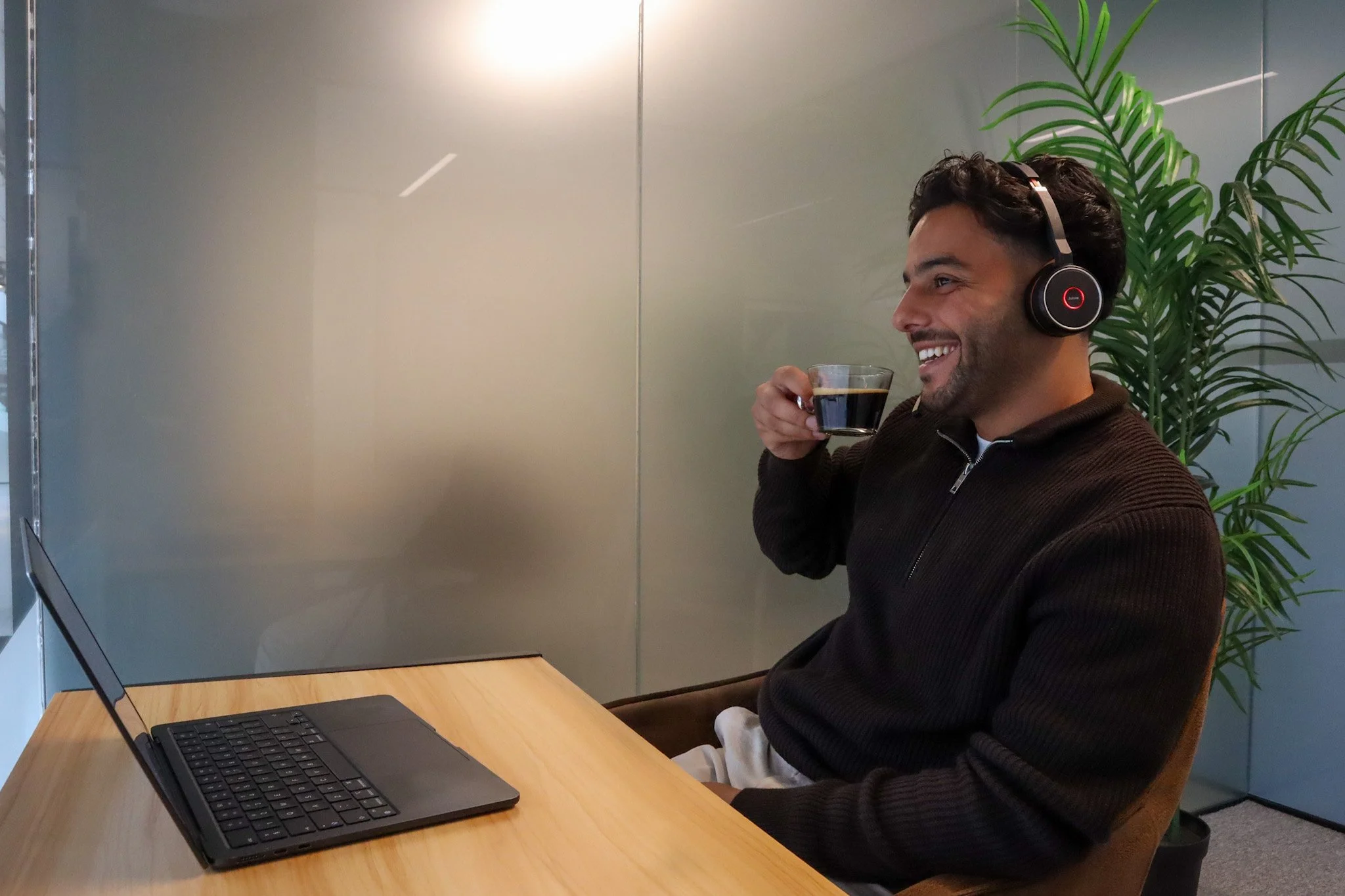 Man with headphones sitting at a desk in a modern office, holding a cup of coffee and smiling, with a laptop in front and a tall green plant behind him.