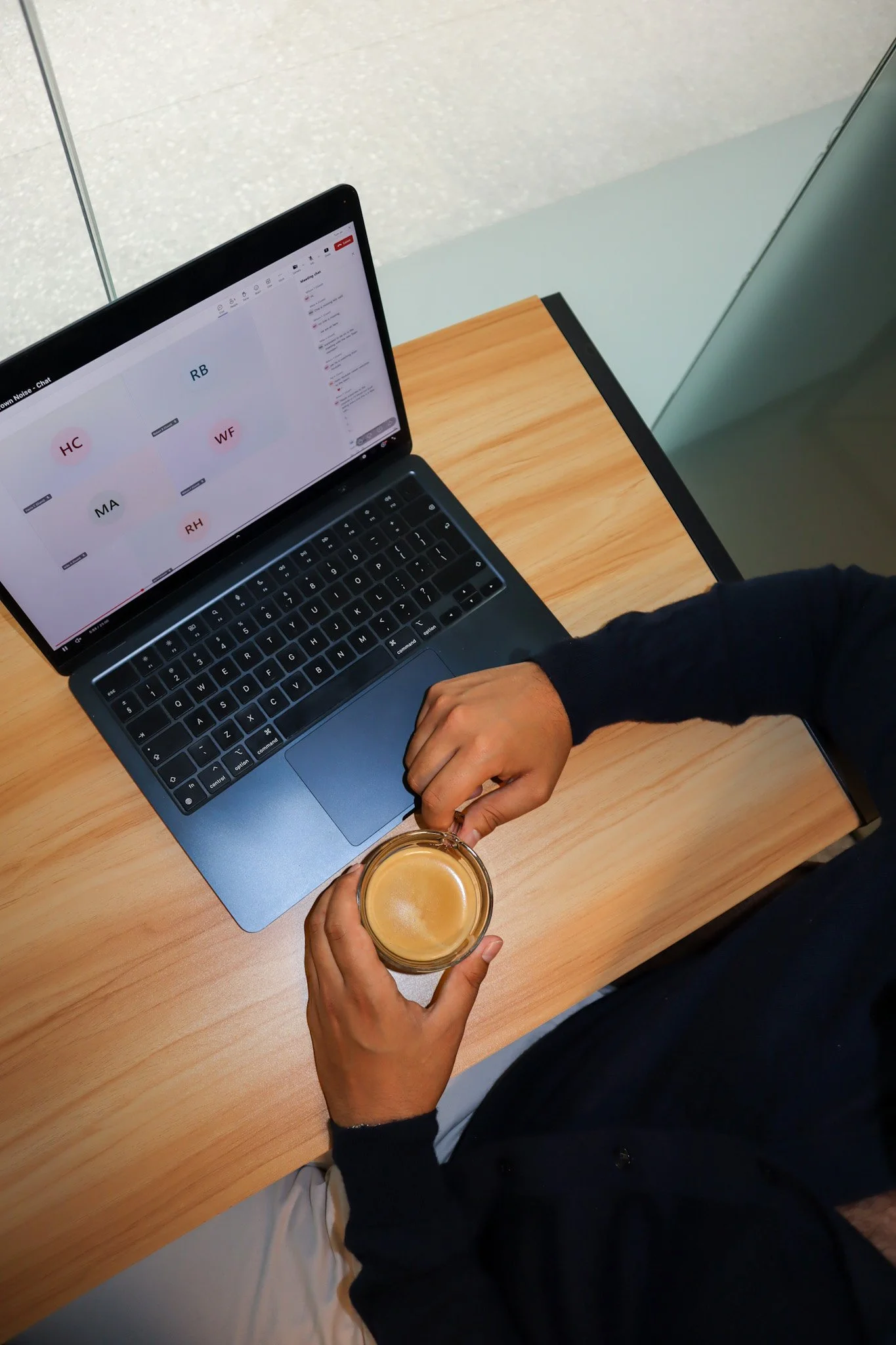 A person sitting at a wooden desk with a laptop, holding a glass of coffee.