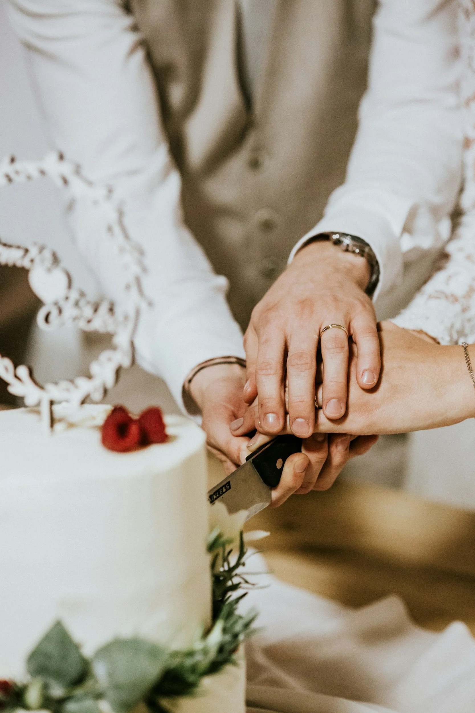 Two people, likely a newlywed couple, are cutting a wedding cake together, holding a knife with multiple hands visible.