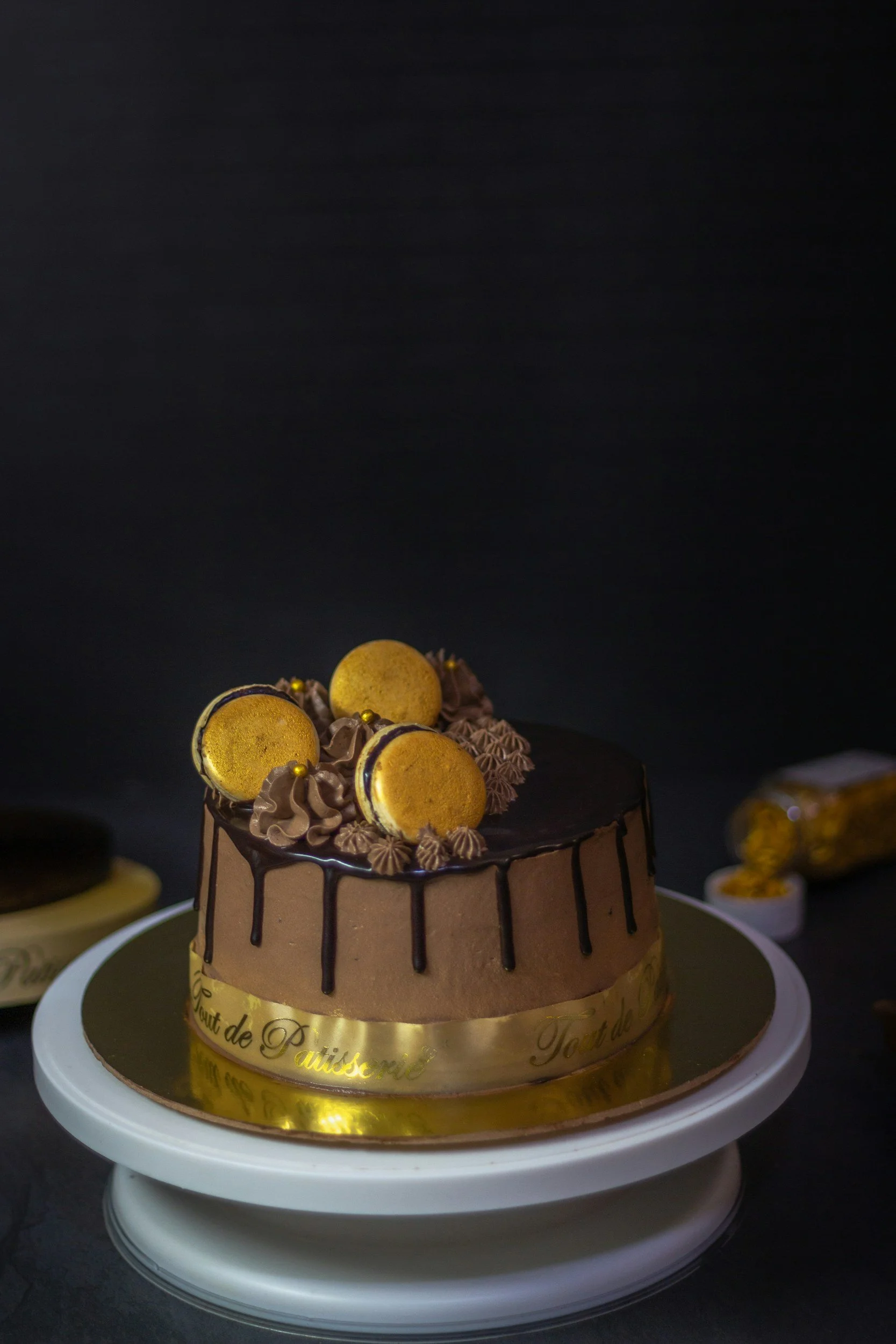 Chocolate cake with citrus macarons and decorations on top, placed on a cake stand with a dark background.
