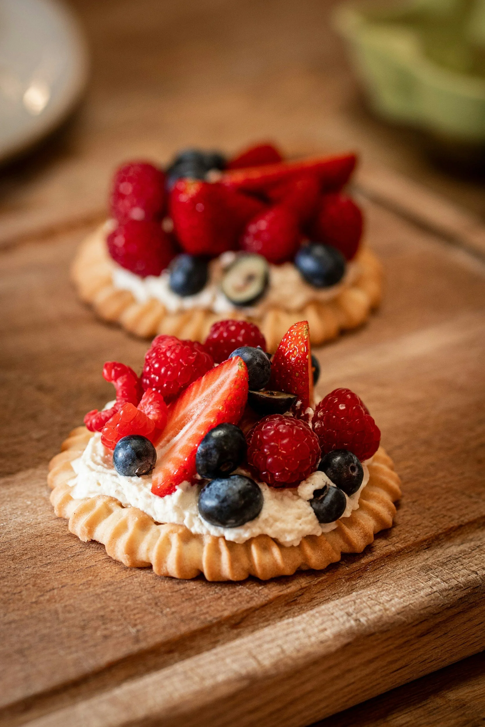 Close-up of two fruit tarts topped with strawberries, blueberries, raspberries, and a small slice of kiwi on a wooden cutting board.