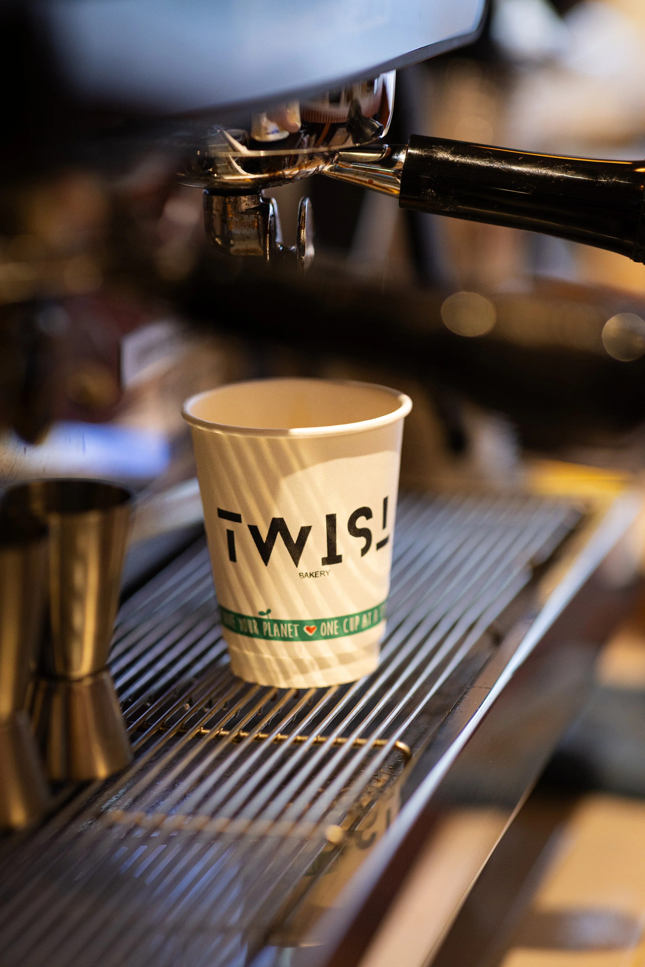 A disposable coffee cup  with the TWIST Bakery & coffee shop logo placed under a coffee machine spout, with the machine in the background.