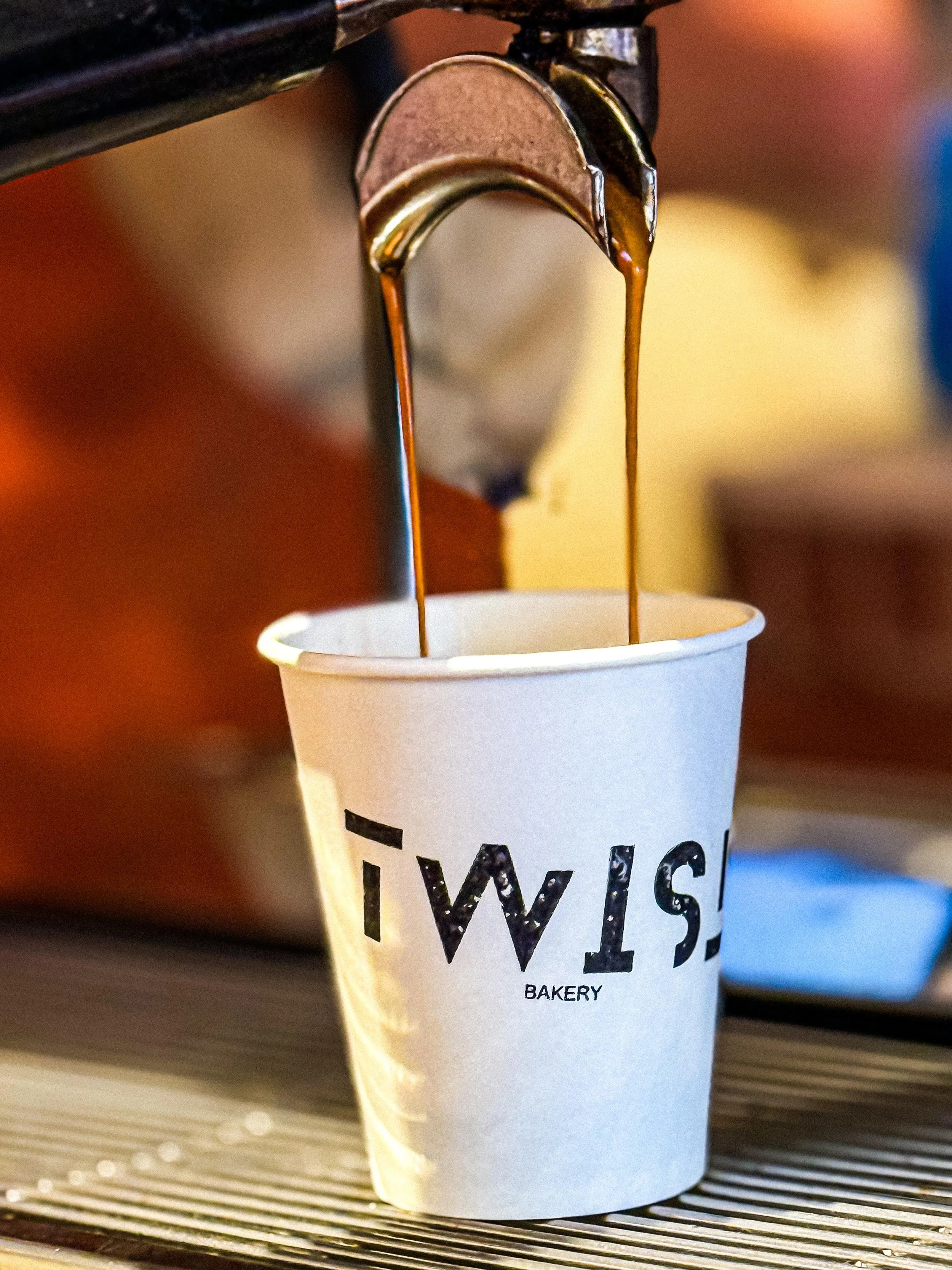 Close-up of a paper cup with the TWIST Bakery & Coffee Shop placed on a coffee machine spout pouring dark coffee into it.