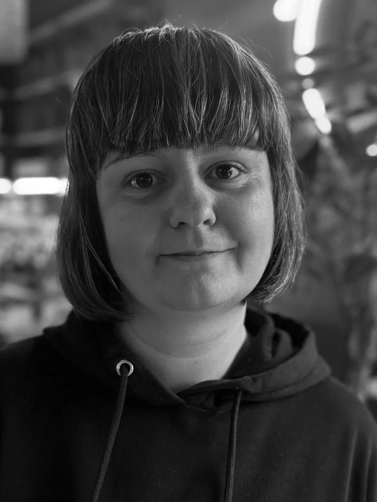 Black and white portrait of a woman with short, straight hair and a slight smile, wearing a hoodie, in an indoor setting with blurred background.