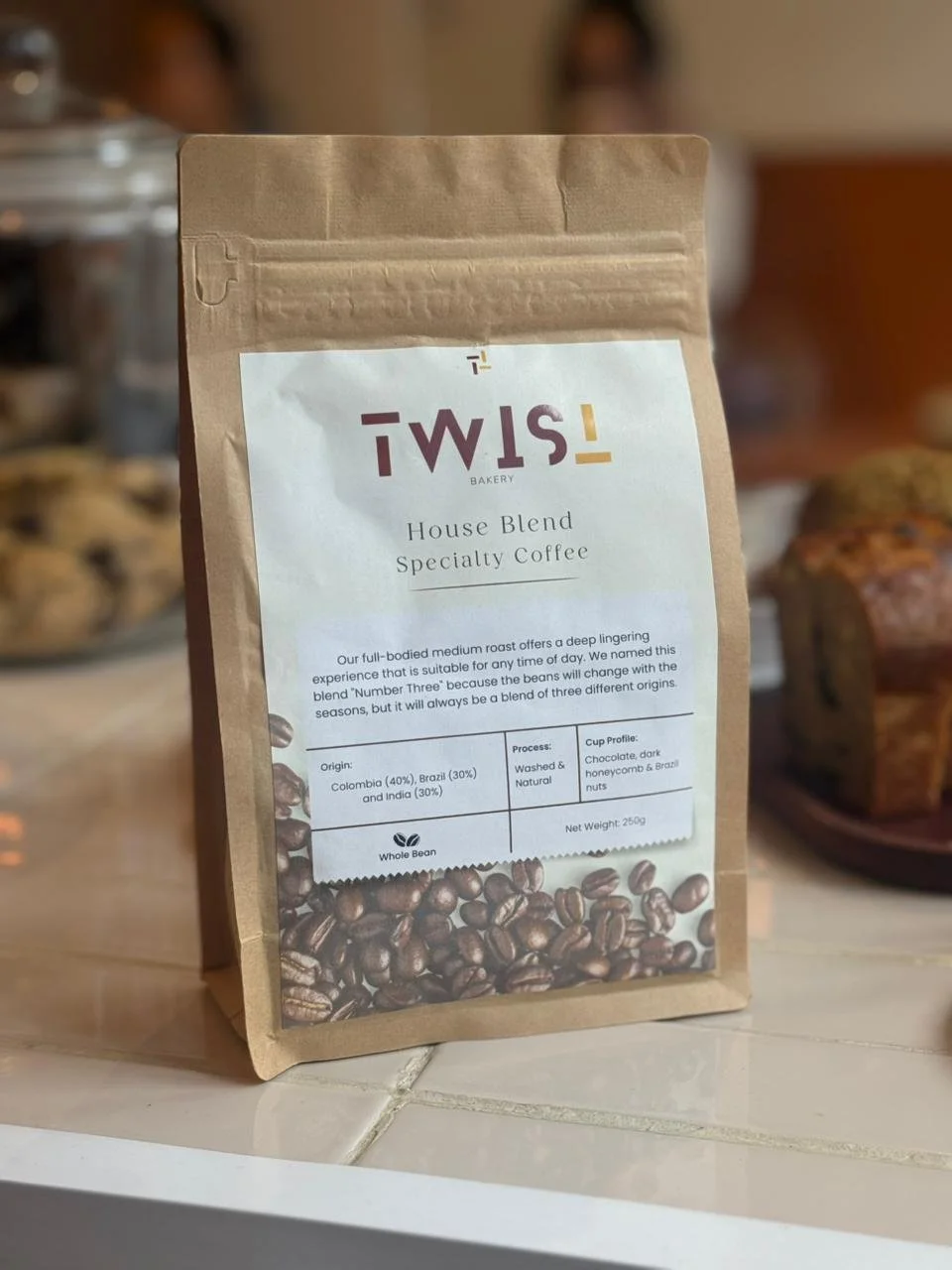 A brown paper bag of Specialty House Blend Coffee from TWIST Bakery & Coffee shop on a countertop, with blurred baked goods in the background.