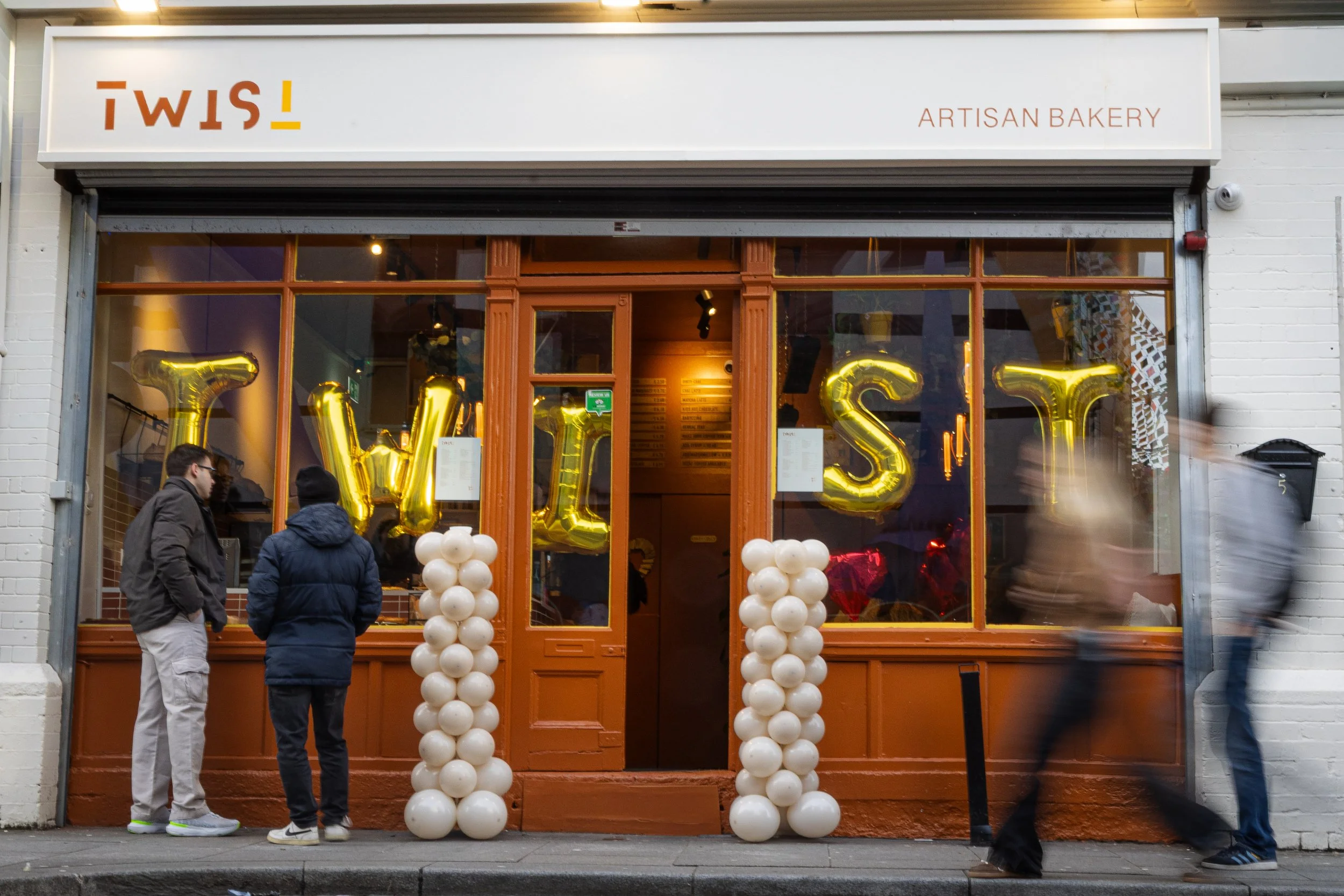 Front exterior of TWIST bakery & coffee shop with large gold balloons spelling out 'TWIST' in the window and white balloon columns at the entrance. Several people are standing outside on the sidewalk.