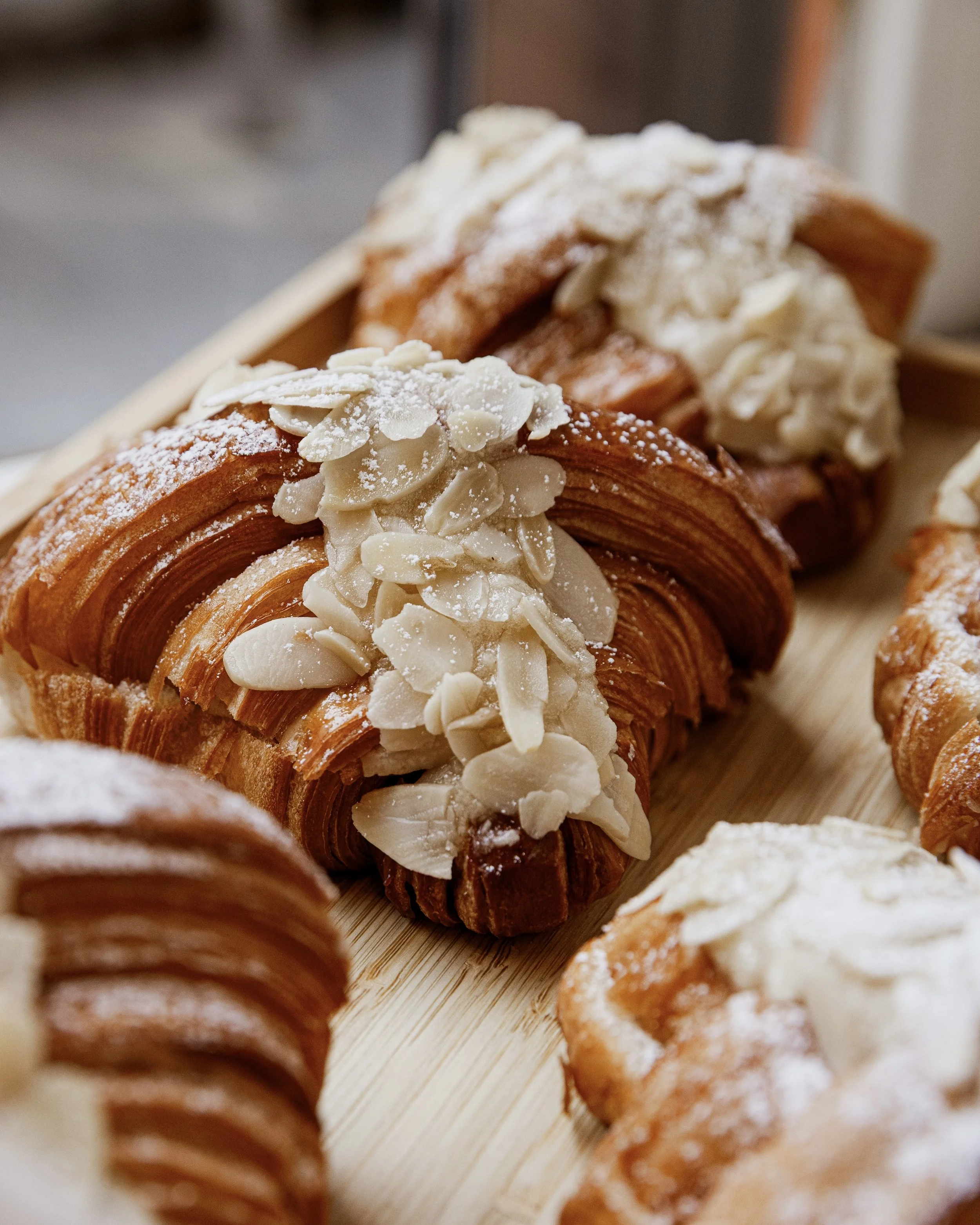Close-up of freshly baked almond croissants from TWIST Bakery dusted with powdered sugar and topped with sliced almonds, served on a wooden board.
