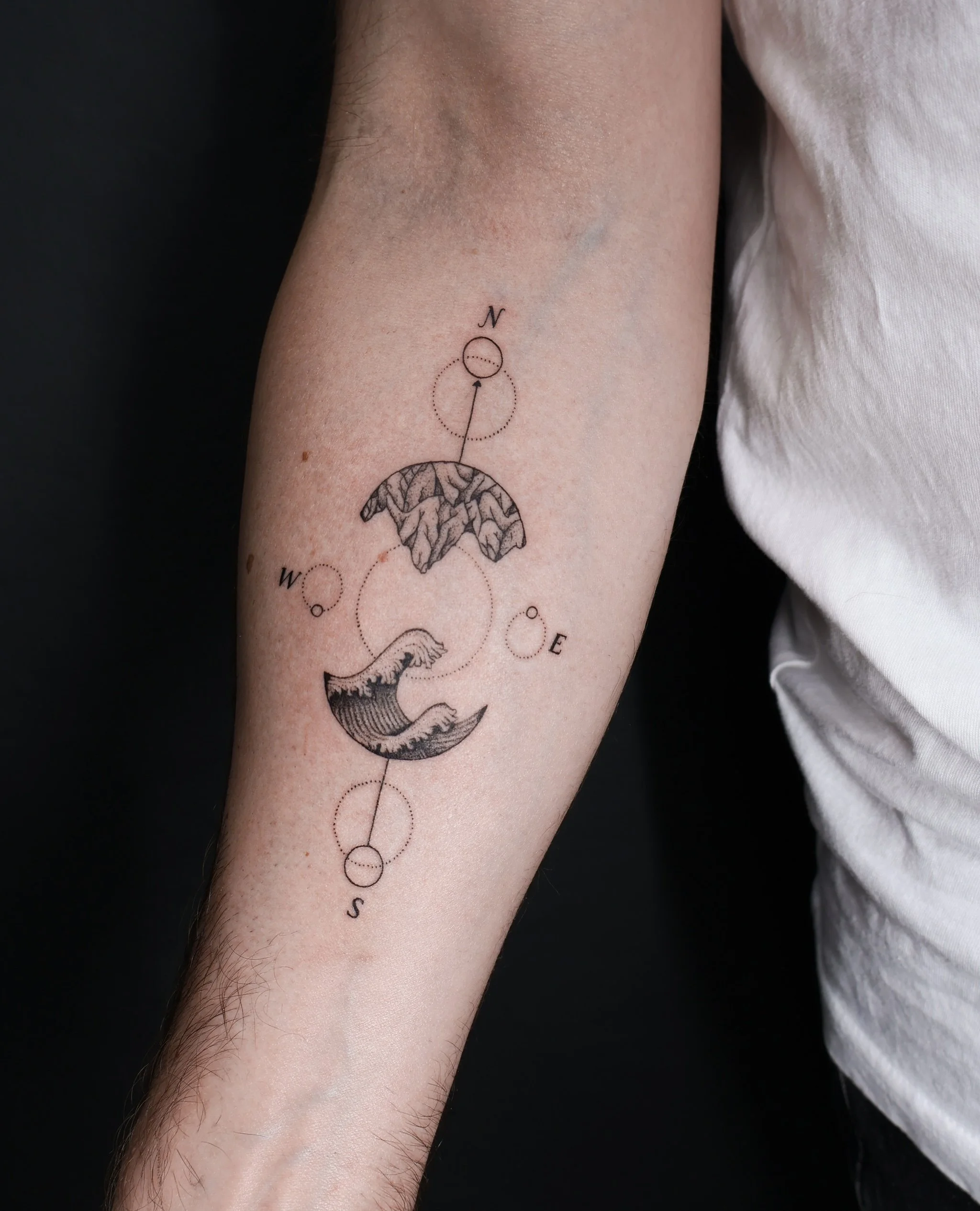 Tattoo on person's inner arm featuring a compass rose with the cardinal directions N, E, S, W, a mountain range, ocean waves, and dotted circle design.