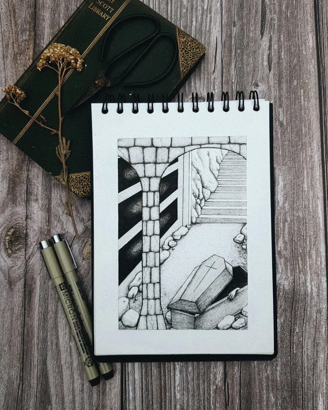 Black and white ink drawing of an outdoor cemetery scene featuring a stone archway, stone stairs, a cross-engraved grave, and rocks, on a spiral-bound sketchpad. Nearby are two black and gold pens, a closed dark green book with gold designs and the t