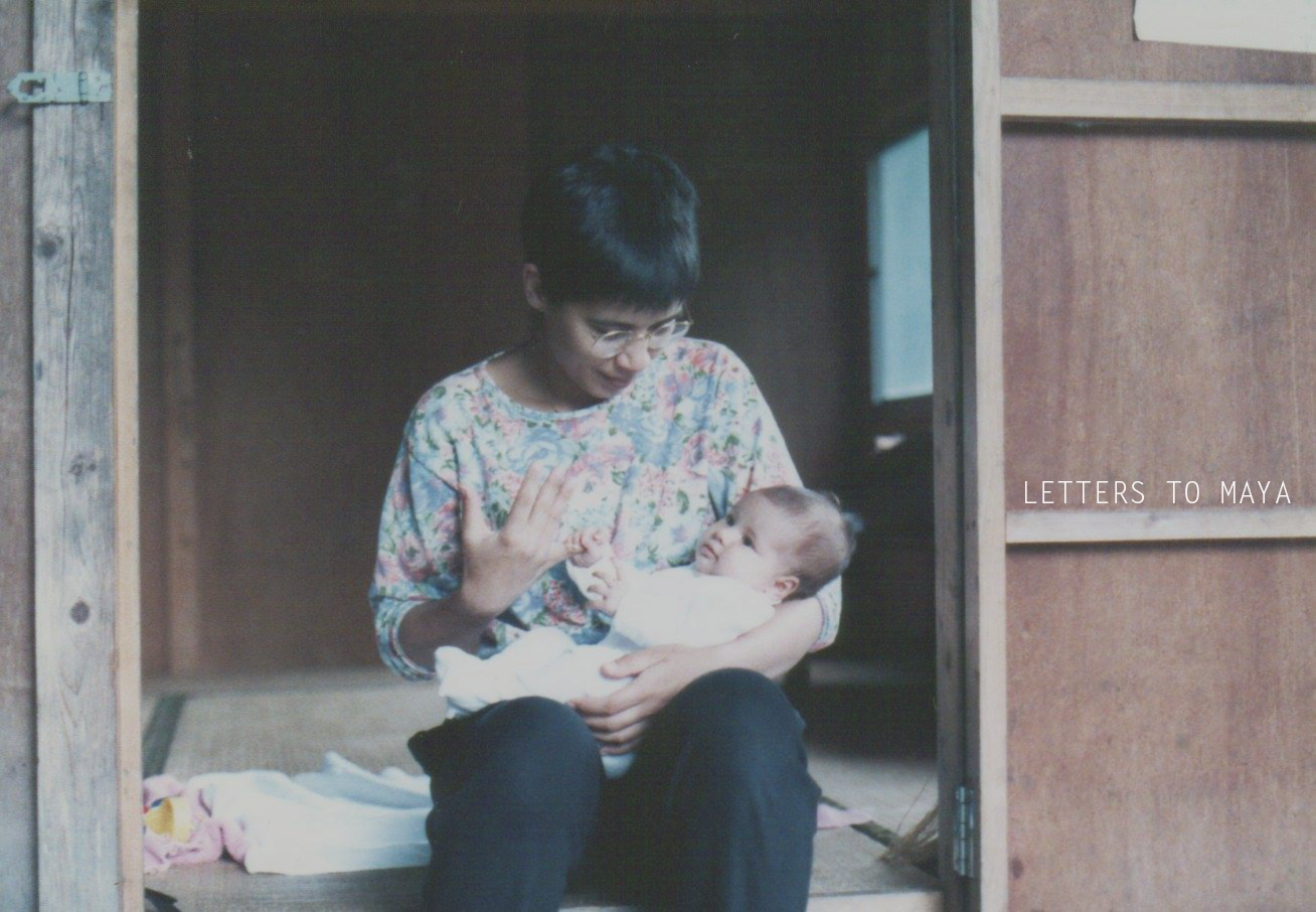 A woman holding a baby and smiling gently, sitting inside a wooden house. The woman is wearing glasses and a colorful floral shirt, and the baby is dressed in white. The scene appears warm and tender.