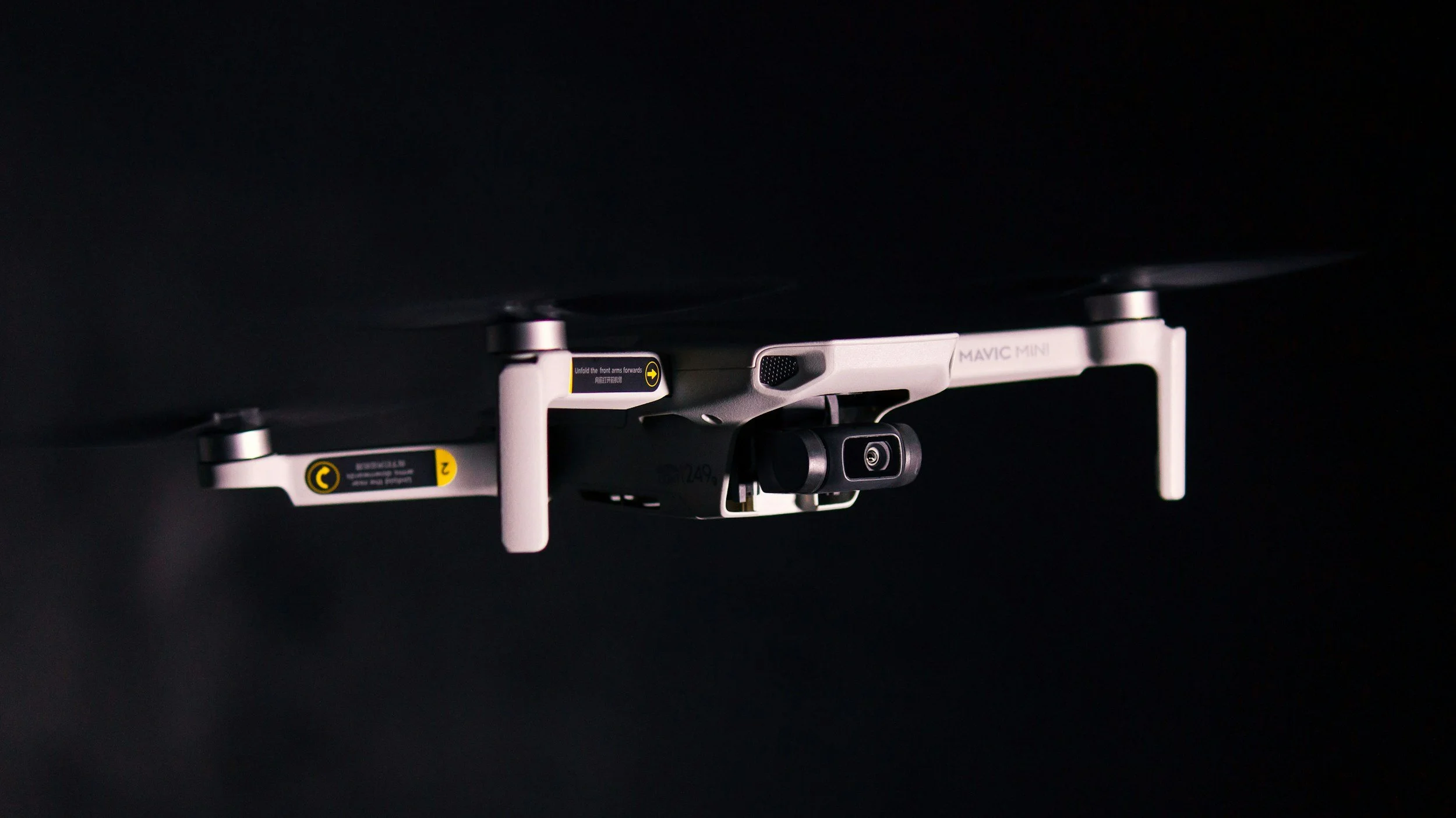 Close-up of a DJI Mavic Mini drone flying in a dark environment, revealing its camera and white body.