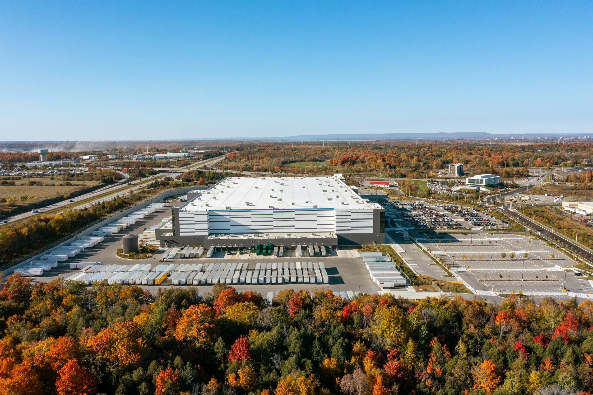 Aerial view of a large warehouse with parking lots and surrounding trees with autumn foliage.