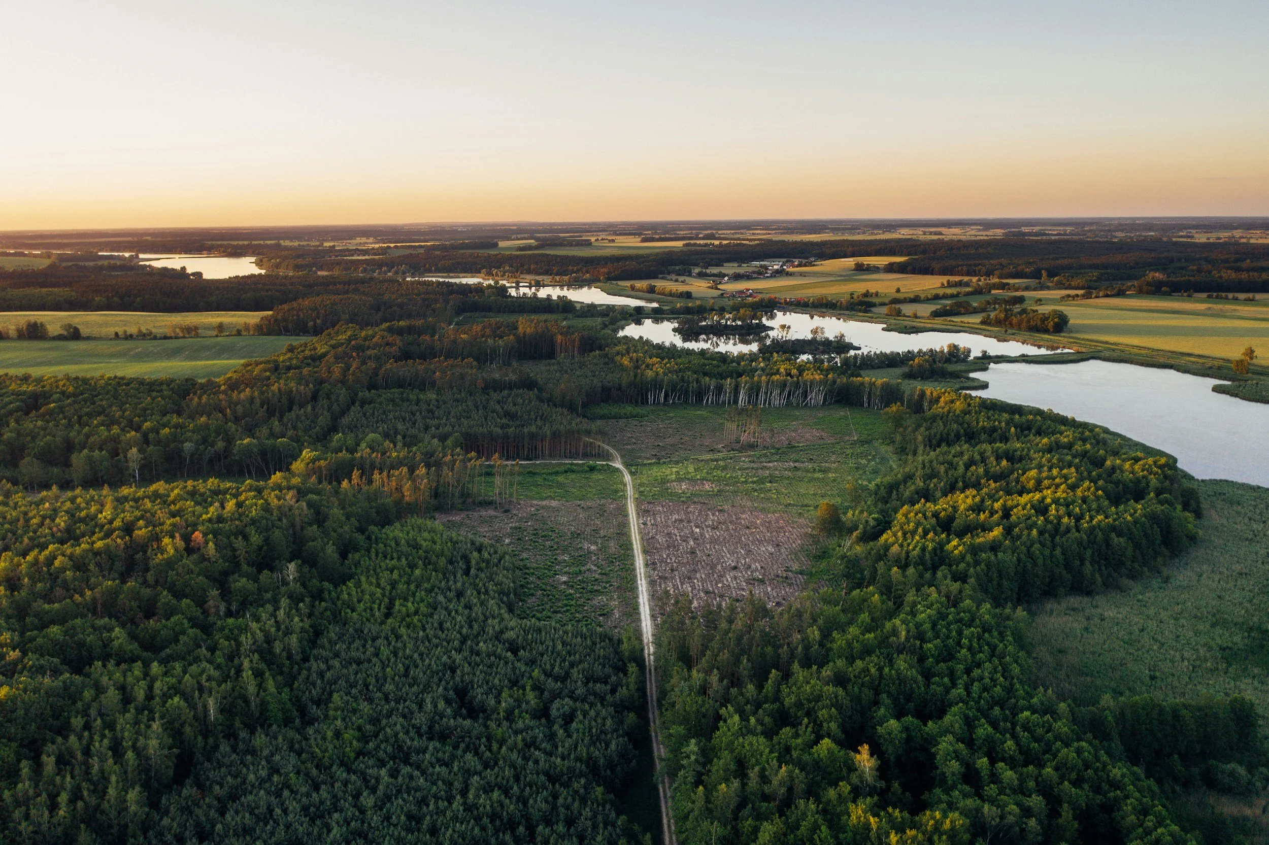 Aerial view of a landscape at sunset featuring lakes, dense forests, open fields, and a dirt road running through the trees.