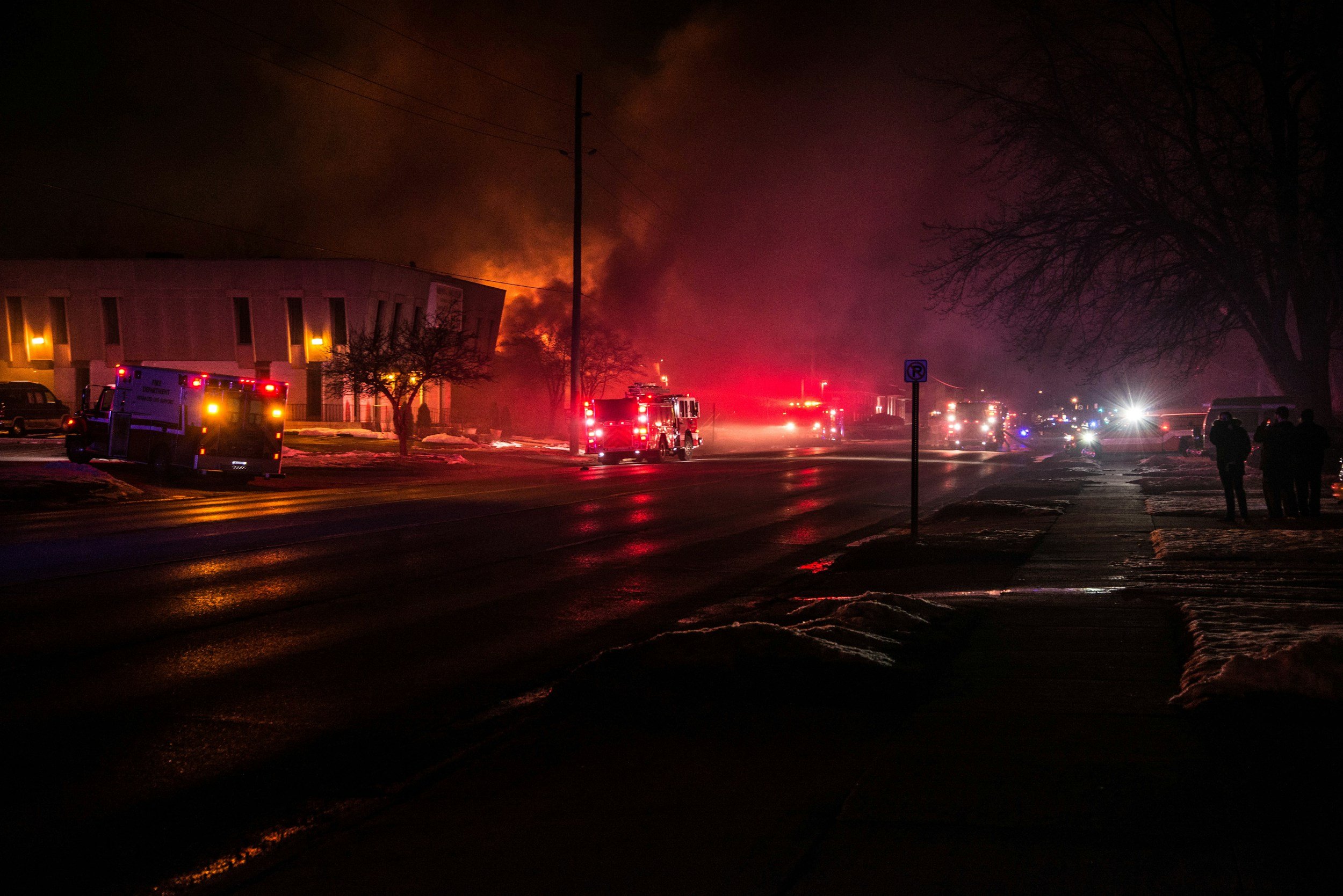 Nighttime scene of a building on fire with thick smoke and flames, emergency vehicles including fire trucks with flashing lights parked on the street, and a few people observing the scene.