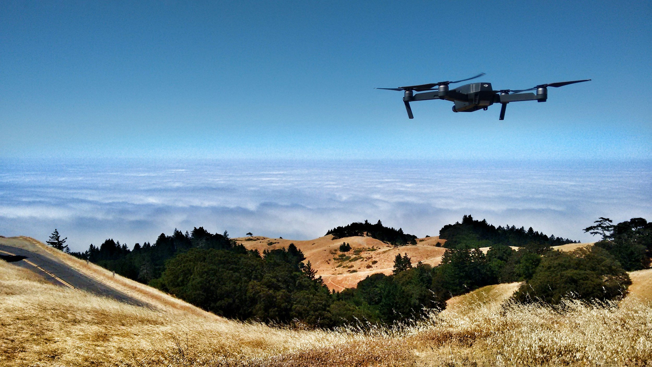 A drone flying over grassy hills with trees, under a blue sky with a layer of clouds in the distance.