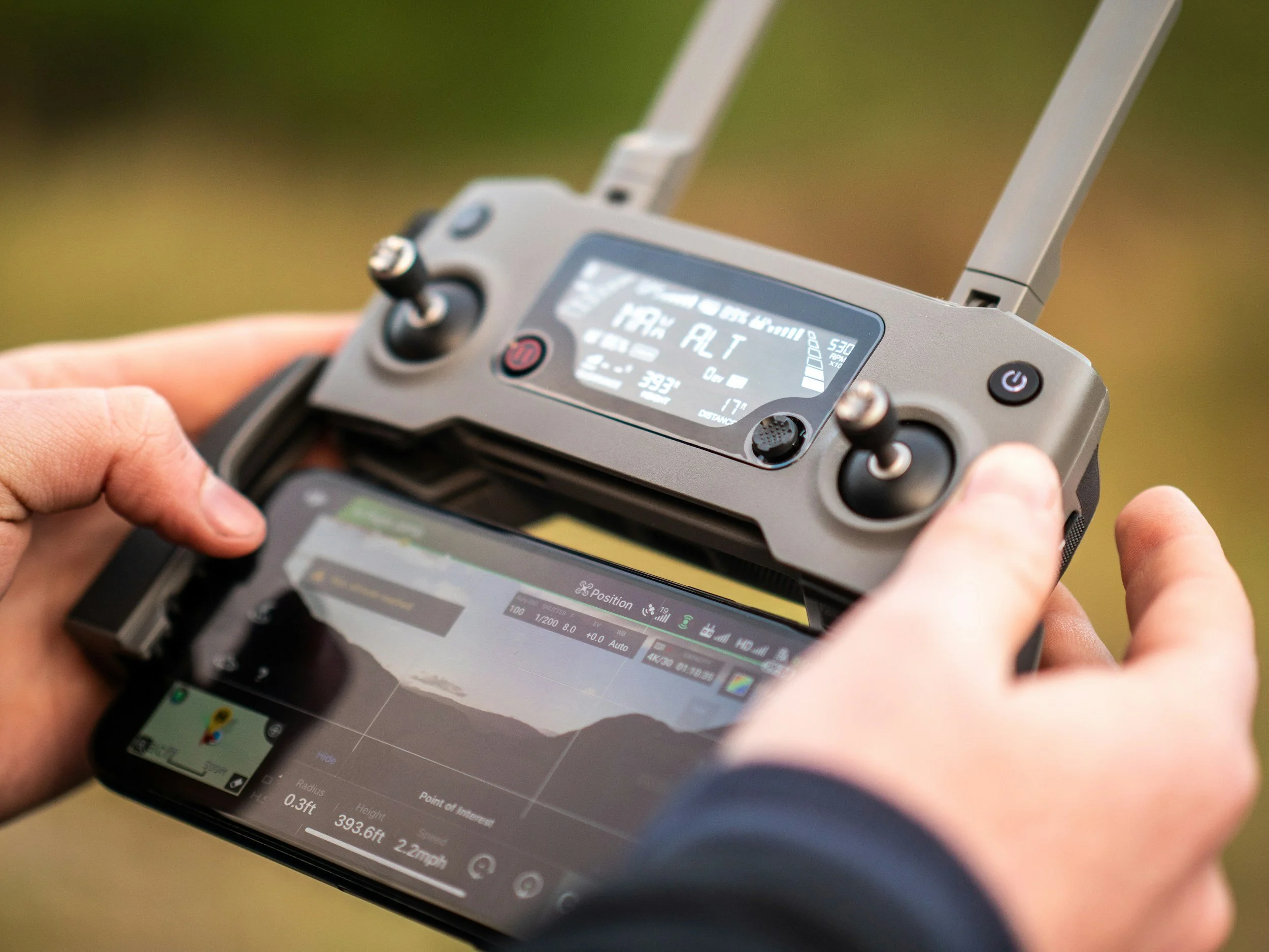 Person operating a drone with a remote controller, which displays altitude, speed, and other data, outdoors during sunset.