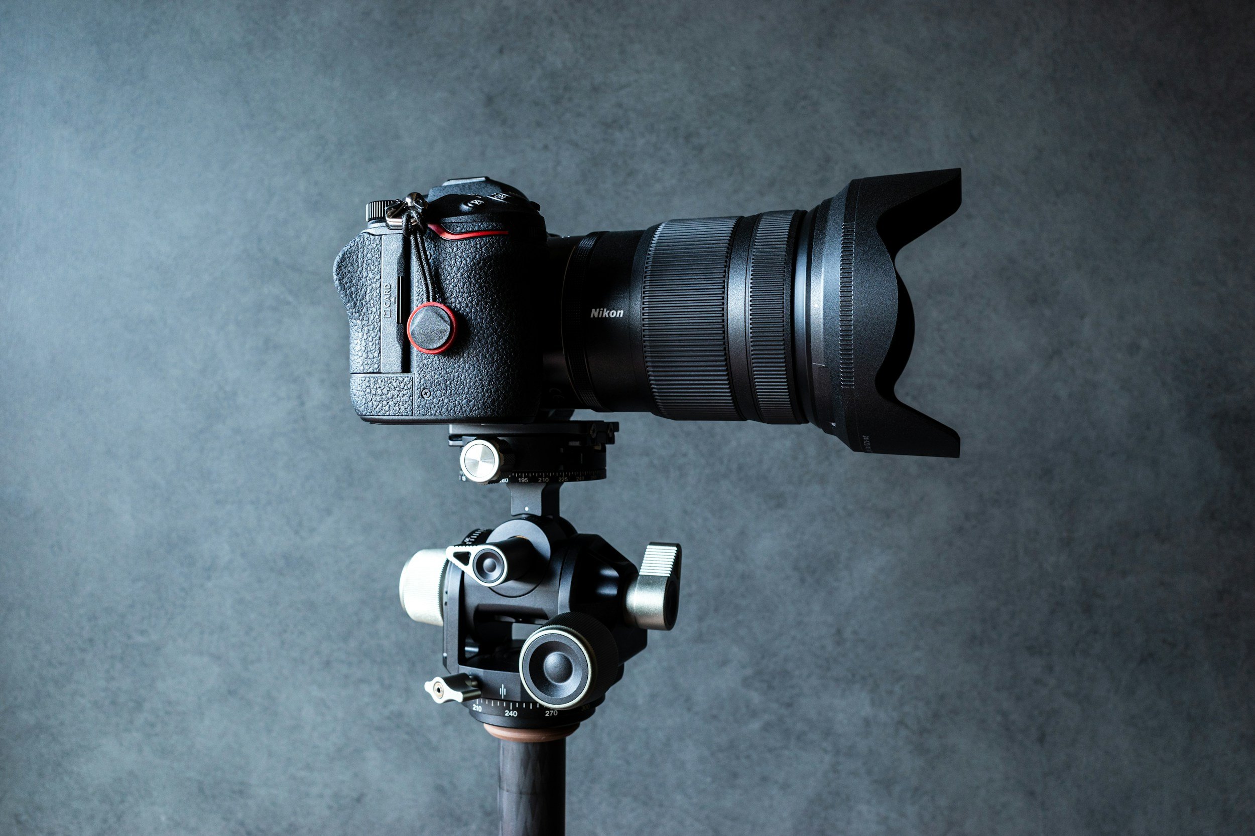 A professional digital camera mounted on a tripod, with a large zoom lens and lens hood, set against a dark textured background.