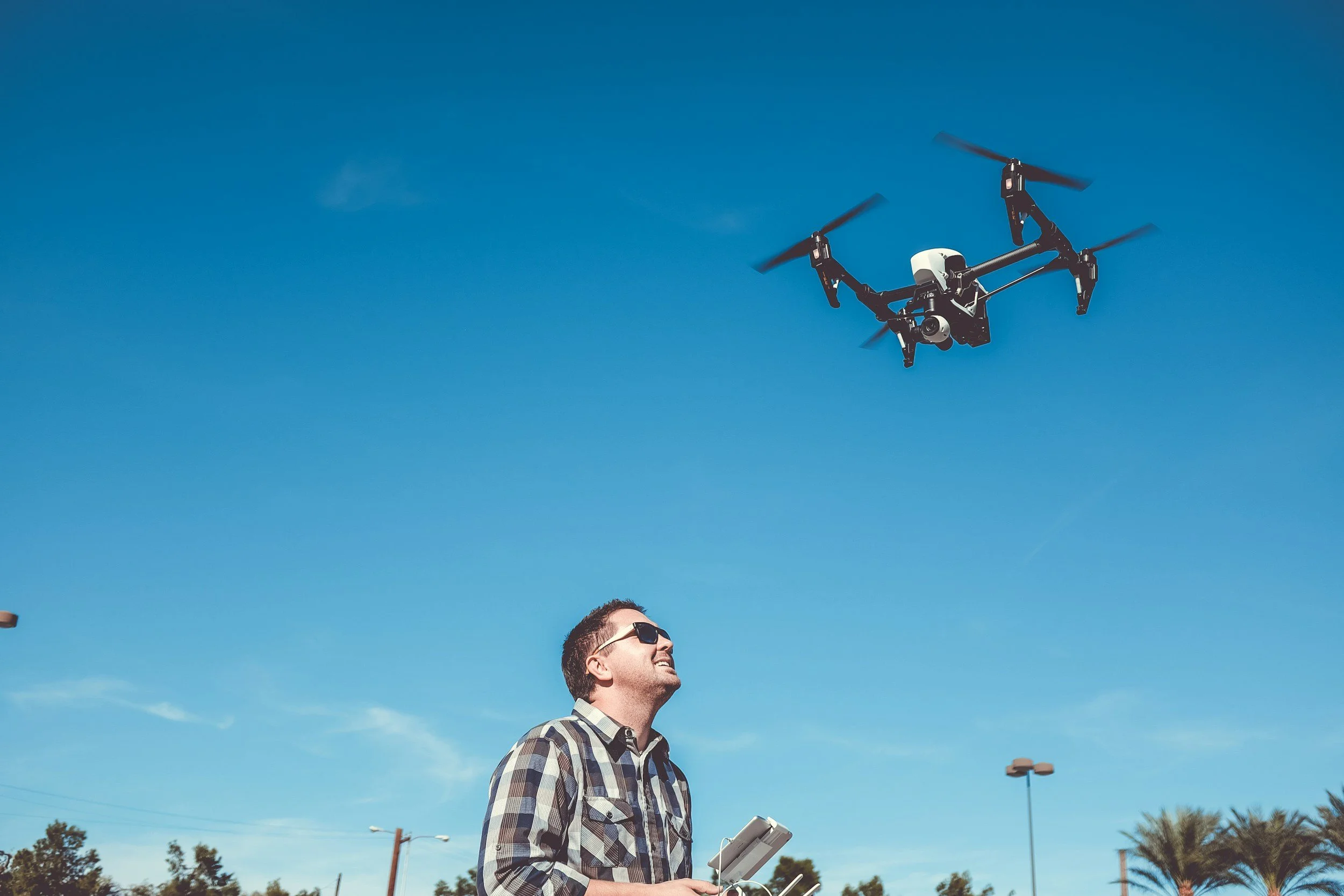 A man flying a drone outdoors on a sunny day, holding a remote control, with palm trees and street lamps in the background.