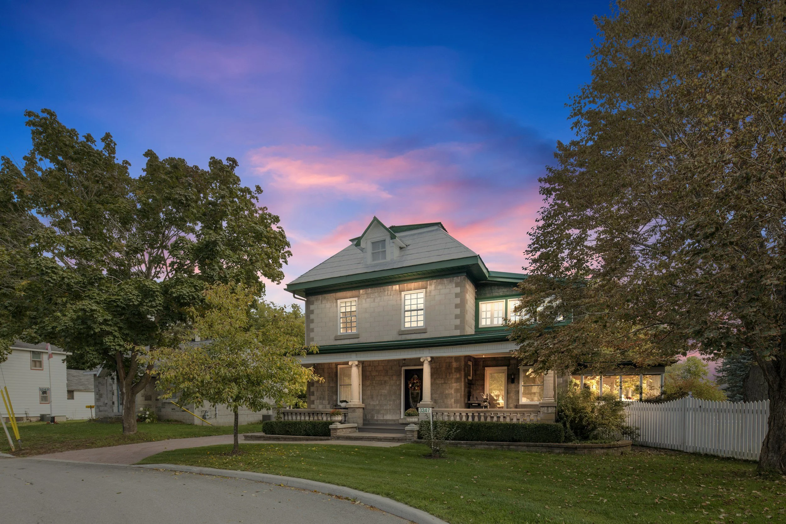 A large, two-story house with a covered front porch, surrounded by trees and a white picket fence. The sky shows pink and blue hues at sunset.
