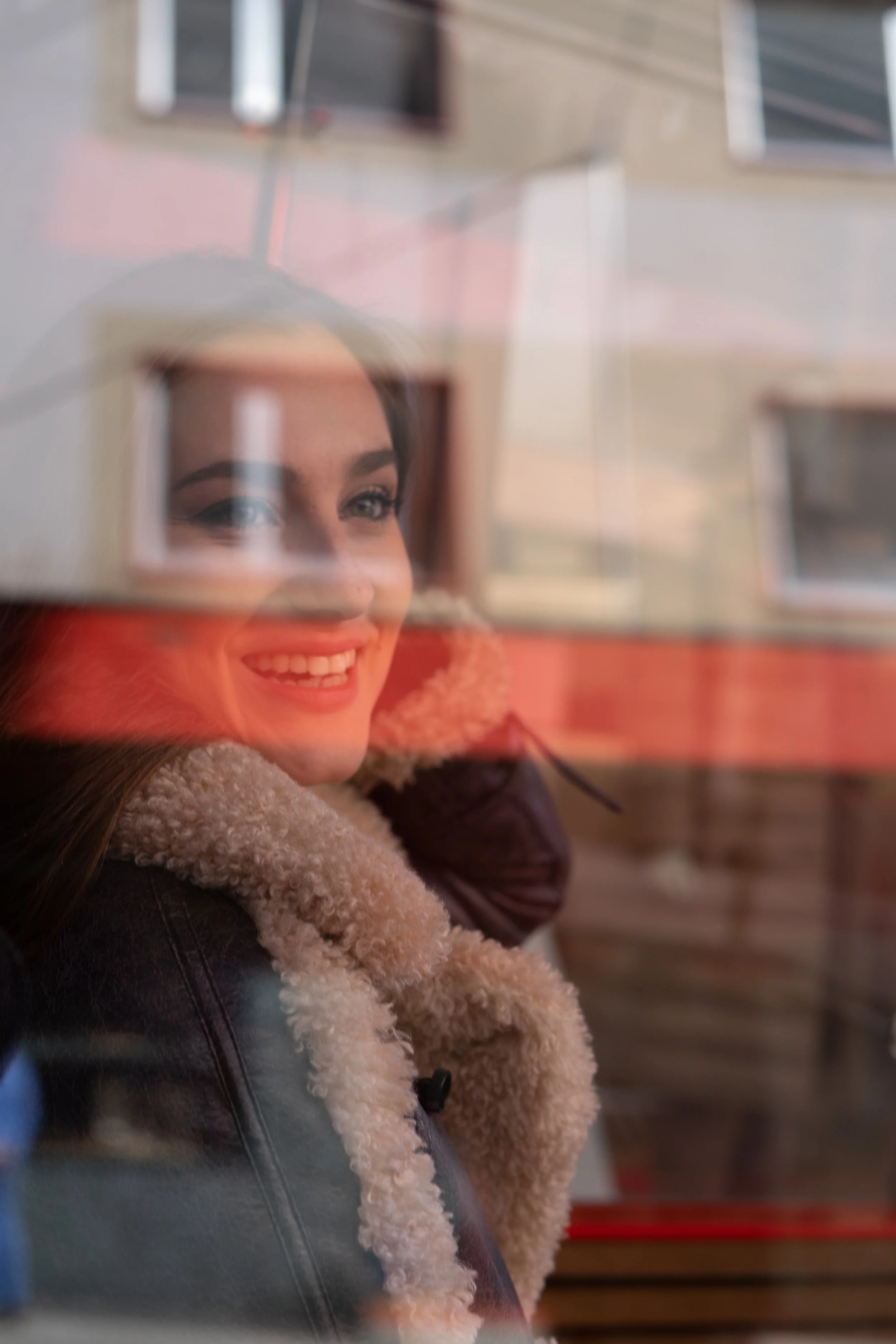 A woman smiling and looking through a glass window, with reflections of buildings visible on the window.