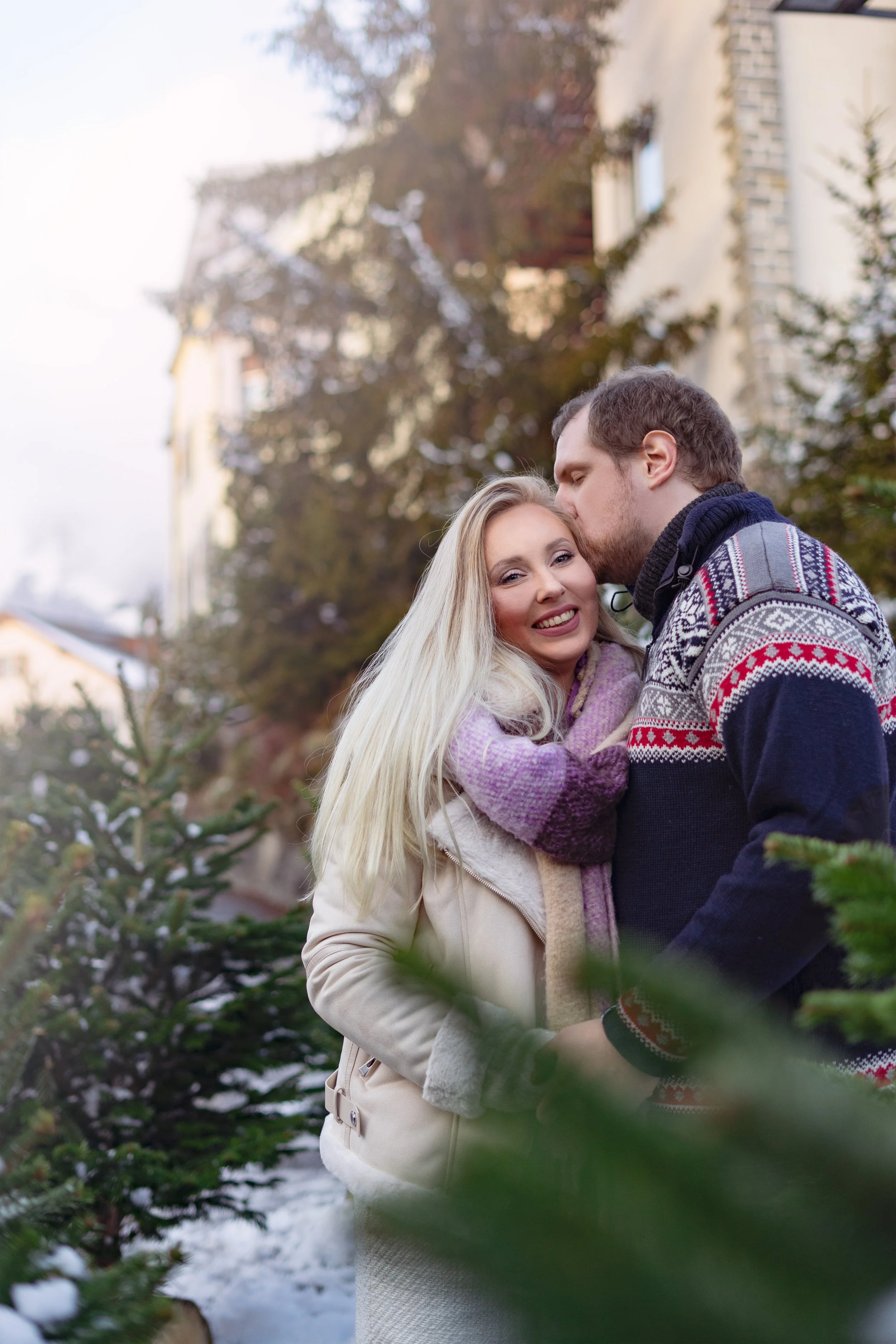 A couple sharing a kiss outdoors during winter, with evergreen trees and snow in the background.