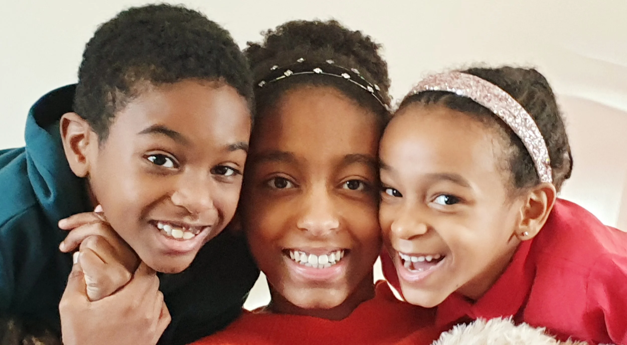 Three smiling children, two girls and one boy, close together facing the camera with cheerful expressions.