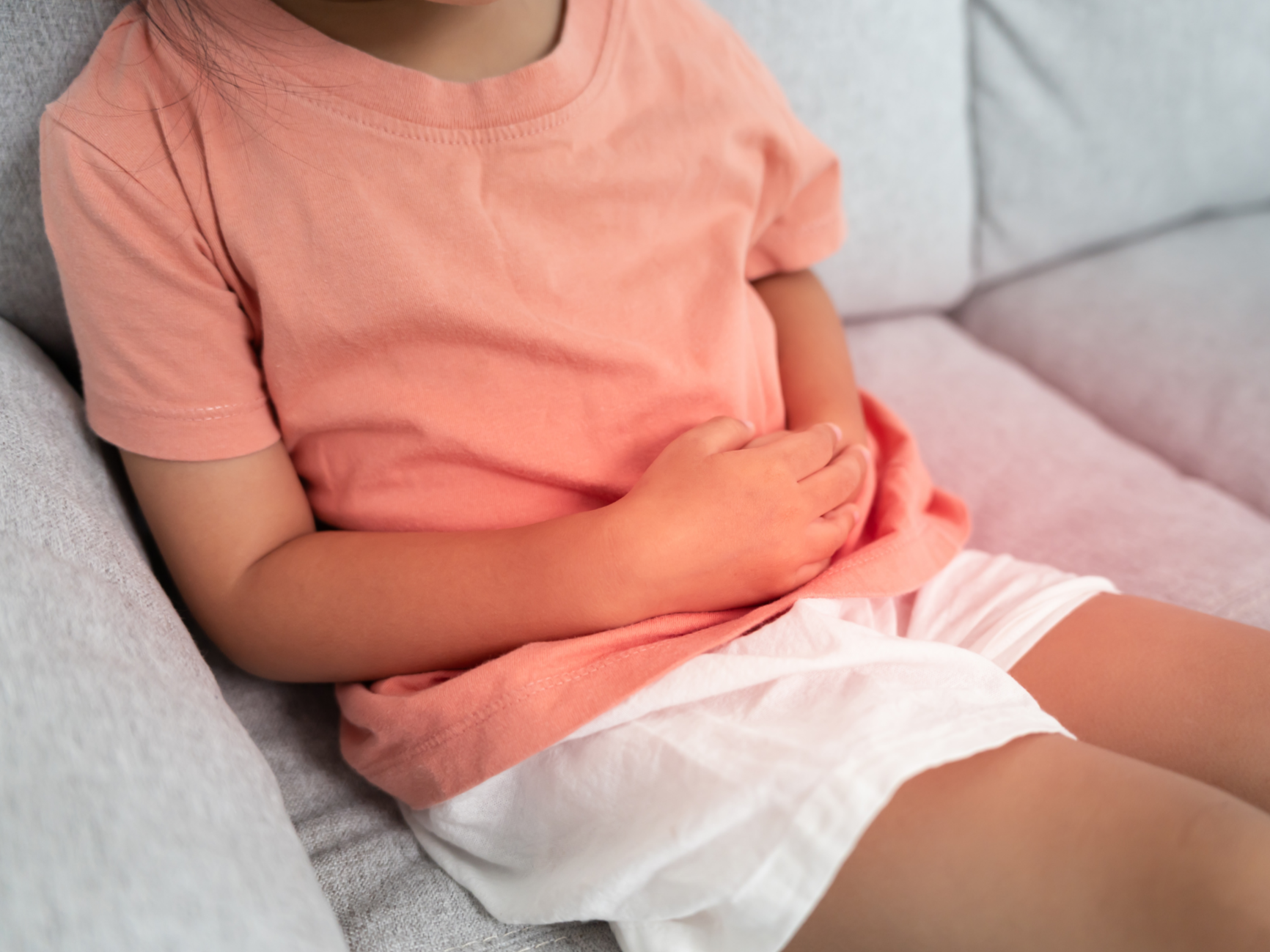 A young girl is sitting on a light gray couch wearing a peach-colored T-shirt and white shorts, holding her stomach with her right hand.