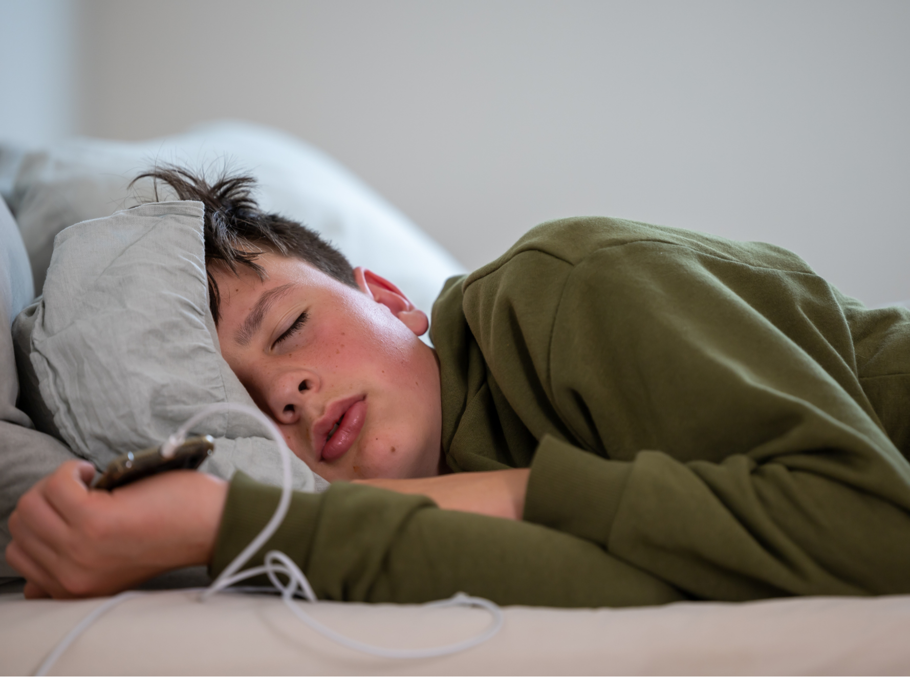 A young person sleeping on a bed, holding a smartphone with earphones plugged in, wearing a green hoodie.