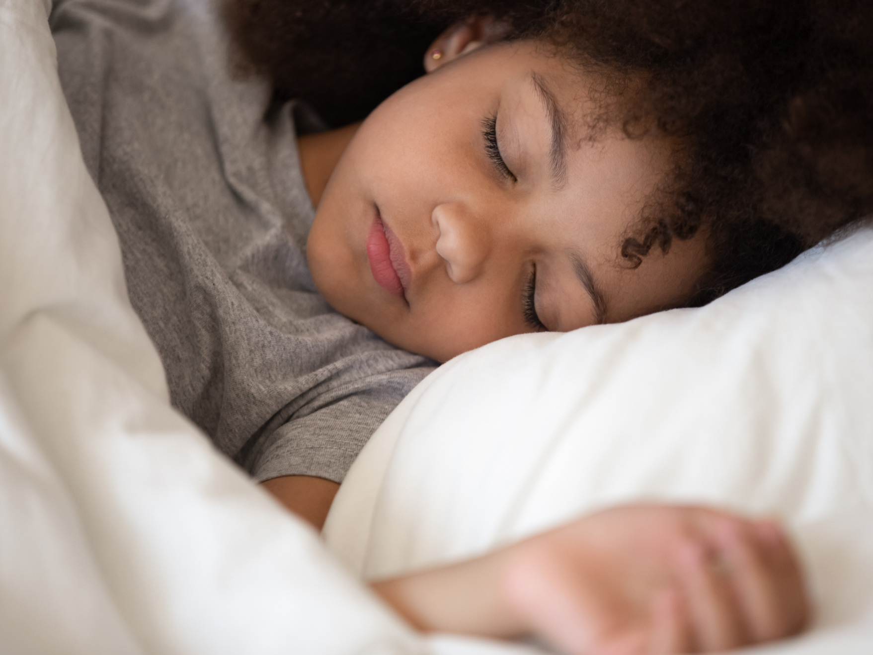 A young woman with curly hair sleeping peacefully on a white pillow, wearing a gray shirt.