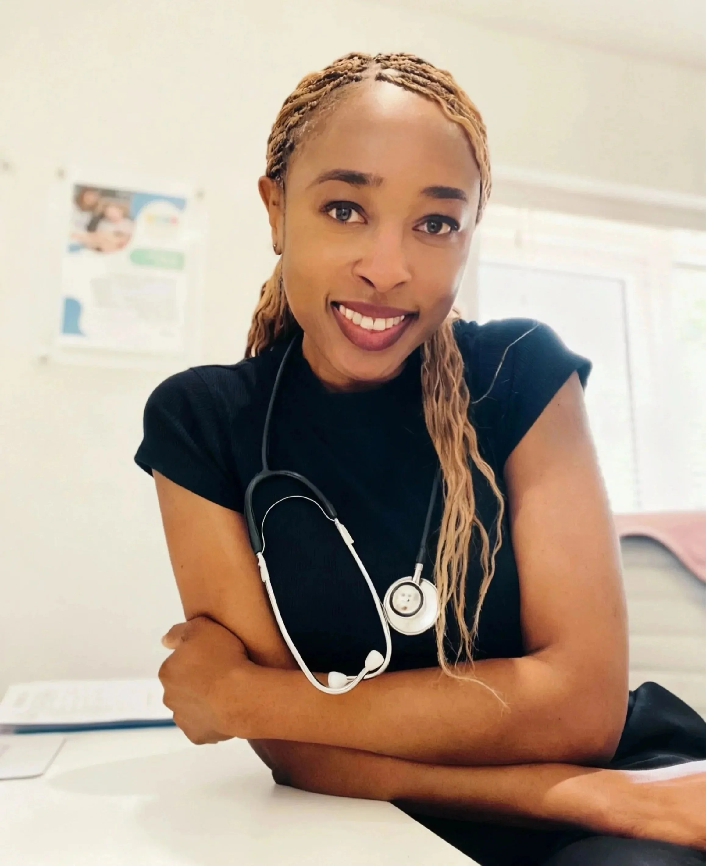 A smiling woman with braids wearing a black shirt and stethoscope around her neck, sitting at a desk in a healthcare setting.