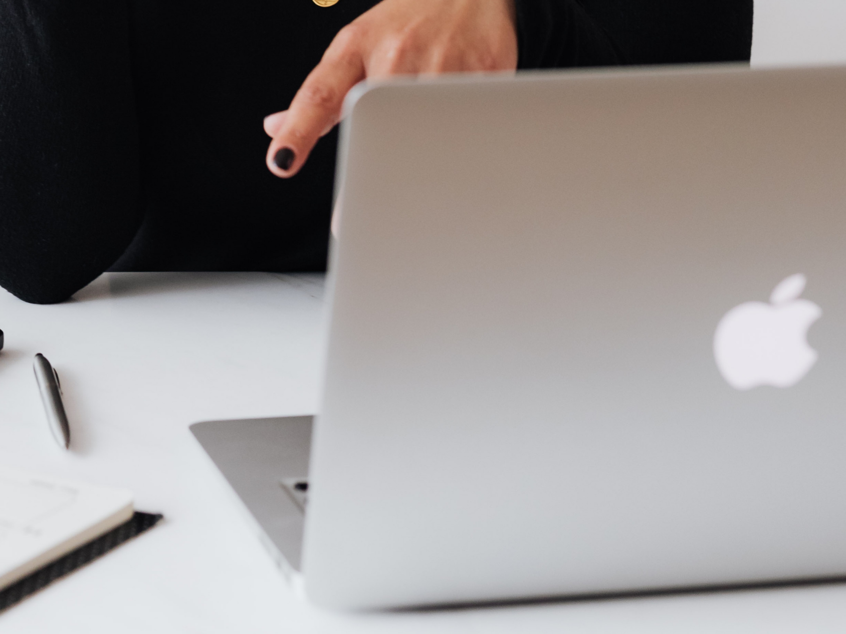 Person in black clothing using a MacBook laptop on white desk, with pen and notepad nearby.