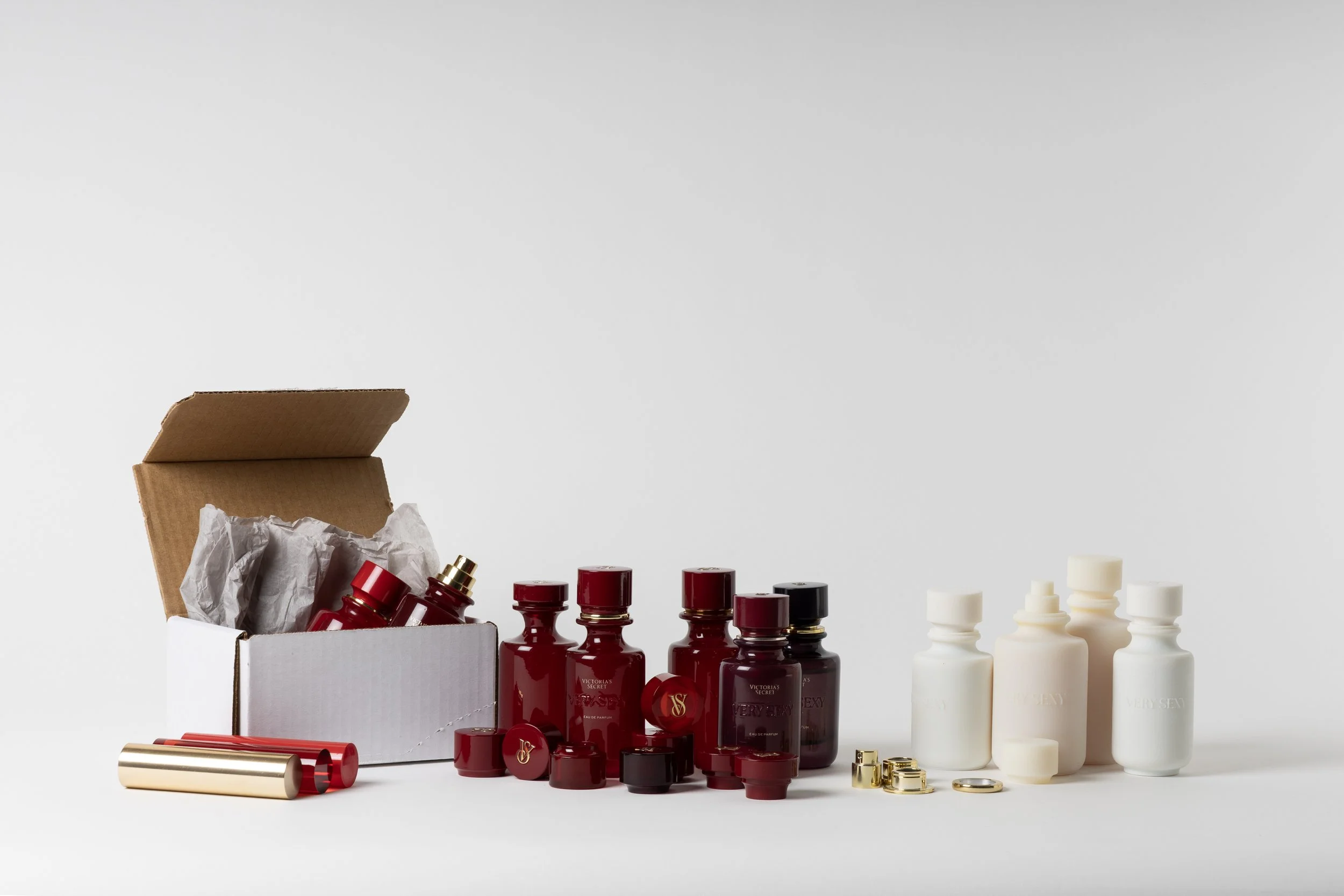 Collection of red and white bottles, jars, and containers, some with gold accents, arranged on a white surface with a cardboard box open in the background.