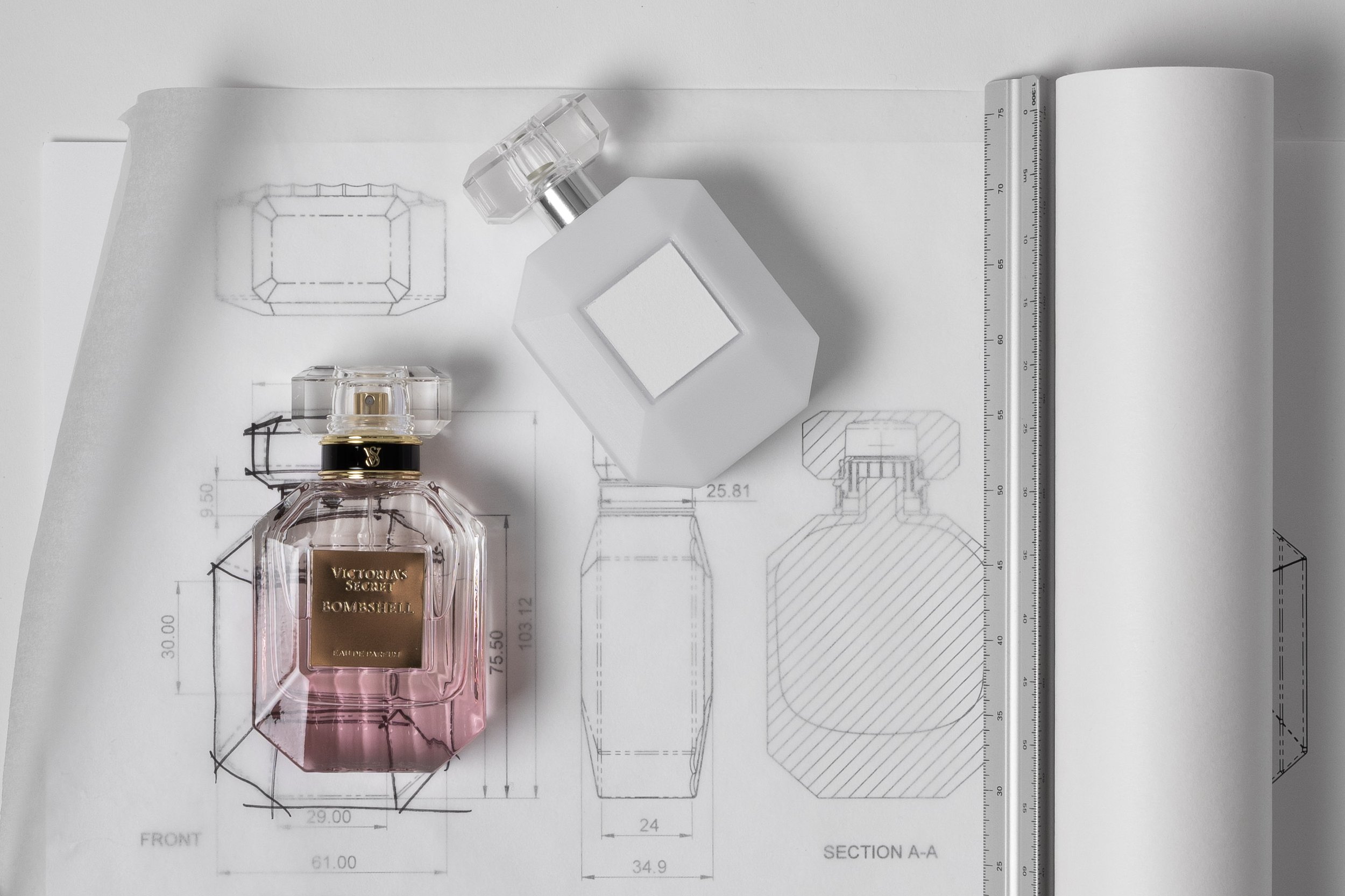 Blueprint of a perfume bottle design with Victoria's Secret Bombshell perfume bottle placed on top, along with technical drawings, measurements, and a ruler.