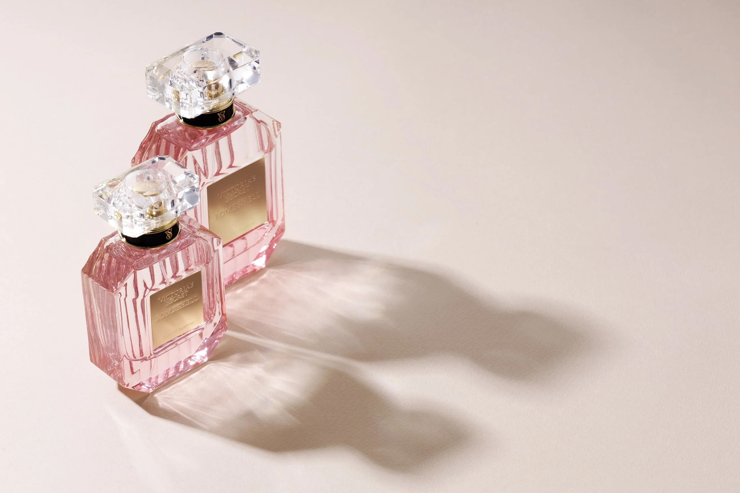 Two Victoria's Secret Bombshell perfume bottles with pink liquid and crystal caps, casting shadows on a plain, light-colored surface.