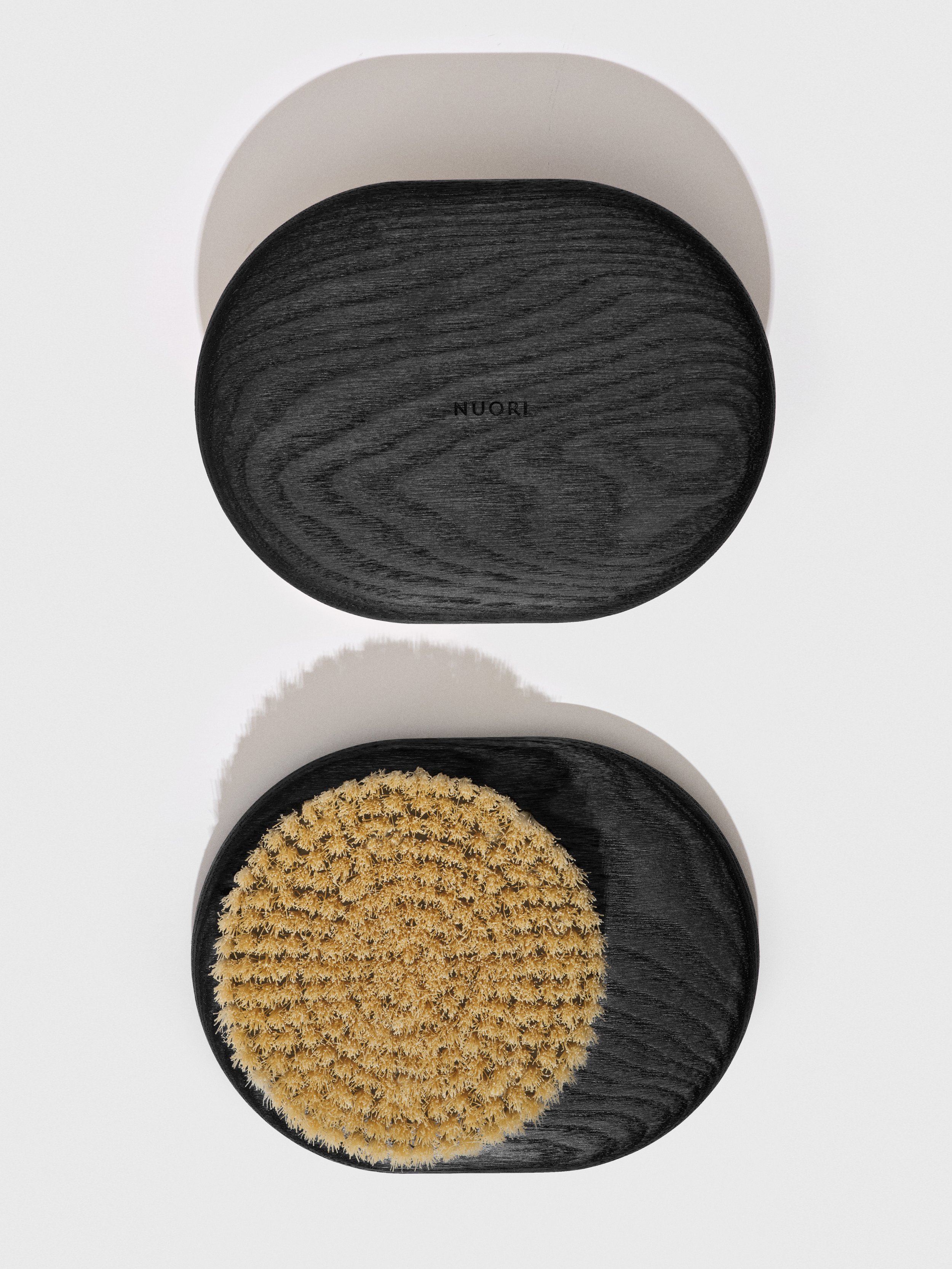 Two black oval-shaped objects, one above the other, with a light-colored background. The top object is solid black with the word 'NUORI' inscribed, and the bottom object has a textured, beige, brush-like surface.