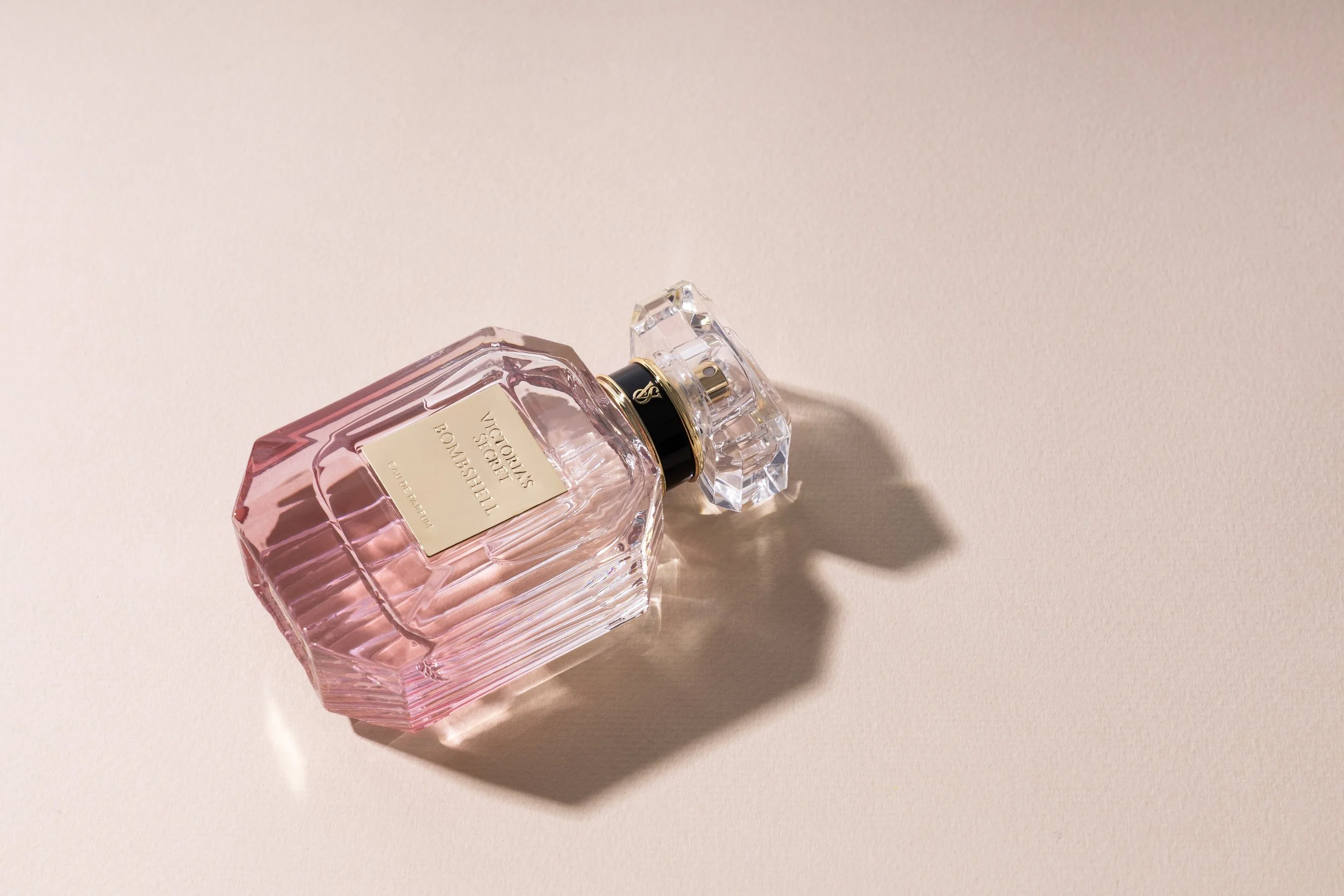 A pink glass perfume bottle with a gold label and a crystal cap on a beige surface, casting a shadow.