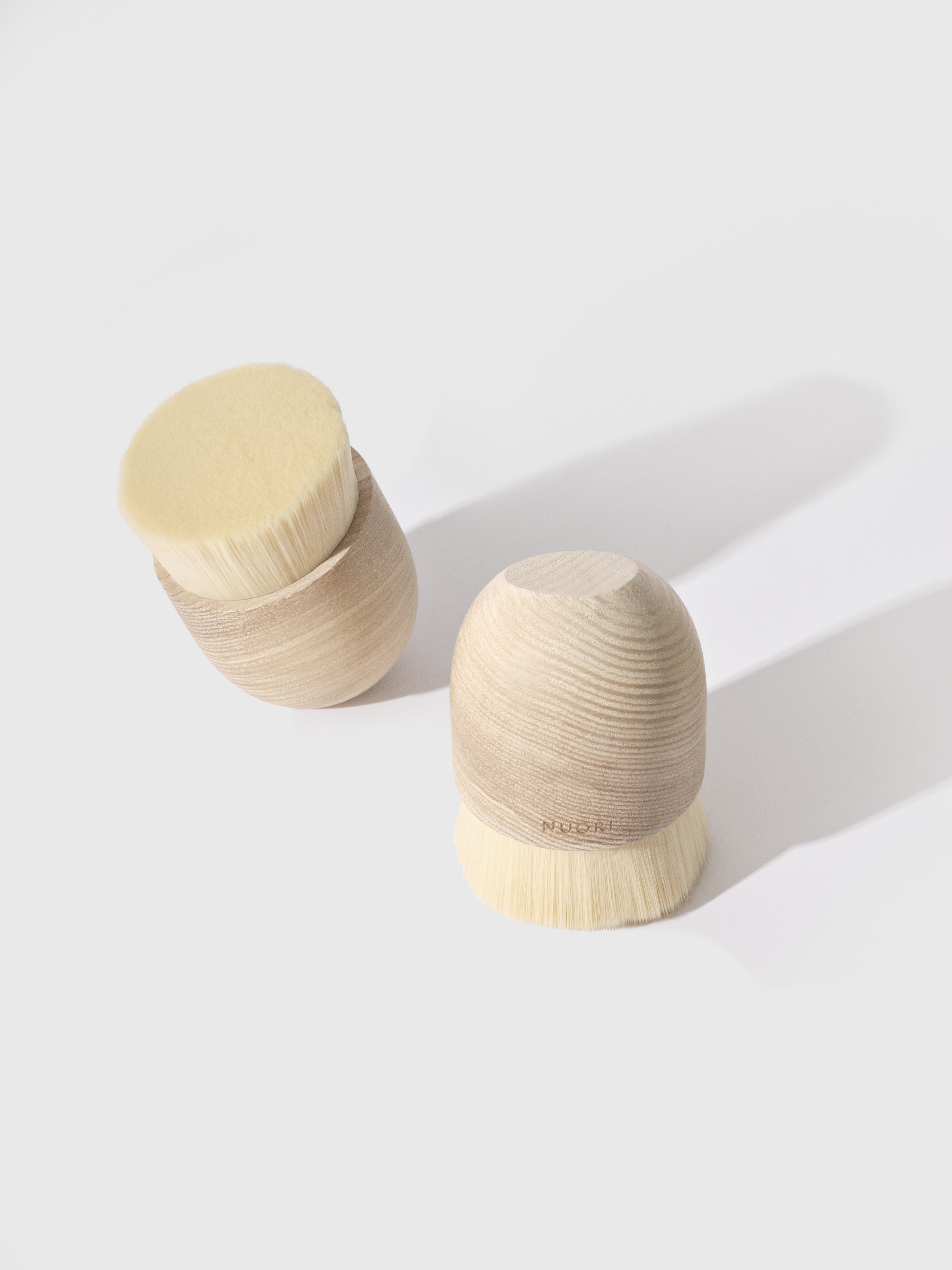 Two wooden brushes with natural bristles, one standing upright and the other lying on its side, on a white surface casting shadows.