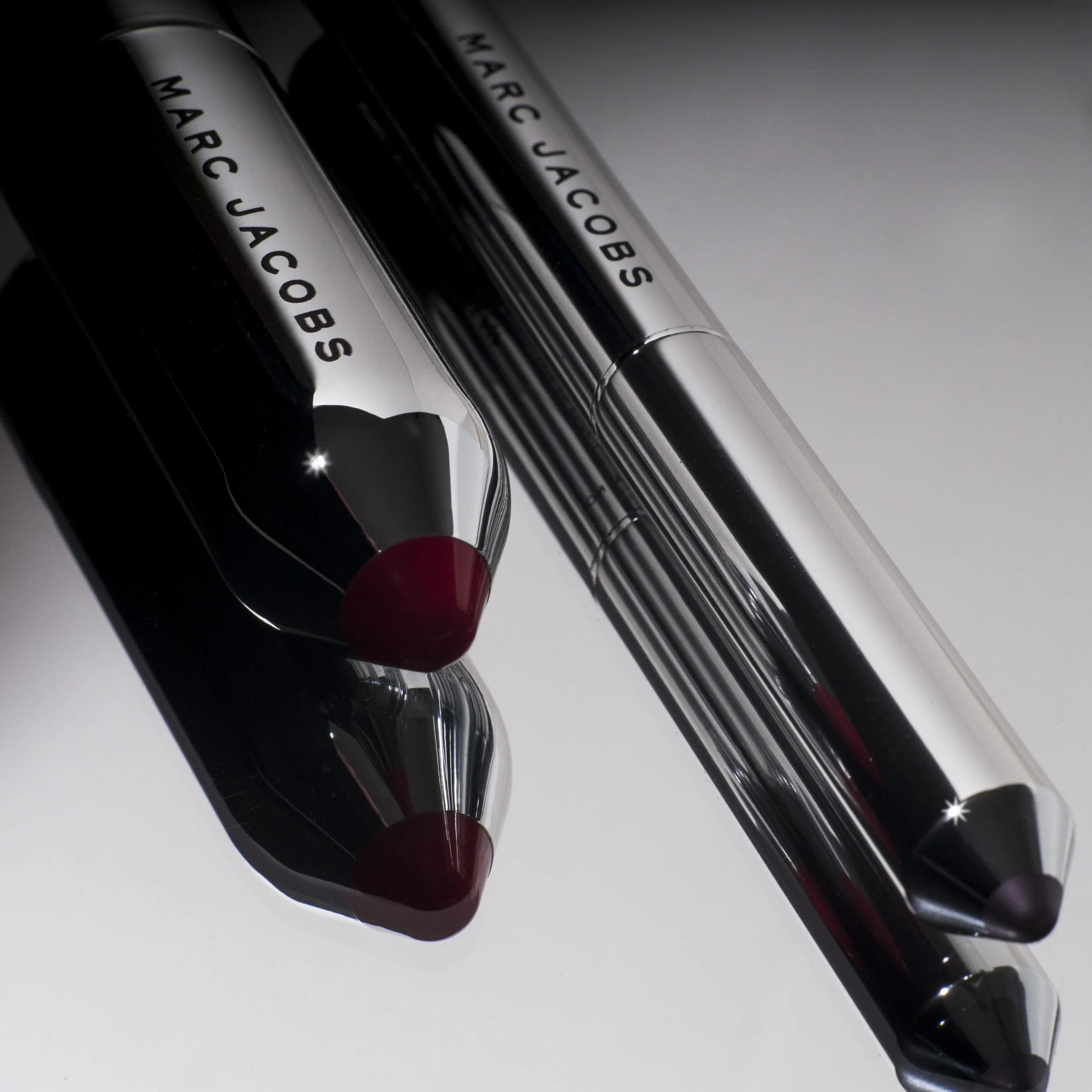 Two Marc Jacobs lip gloss tubes with metallic caps lying on a white surface, reflecting on it.