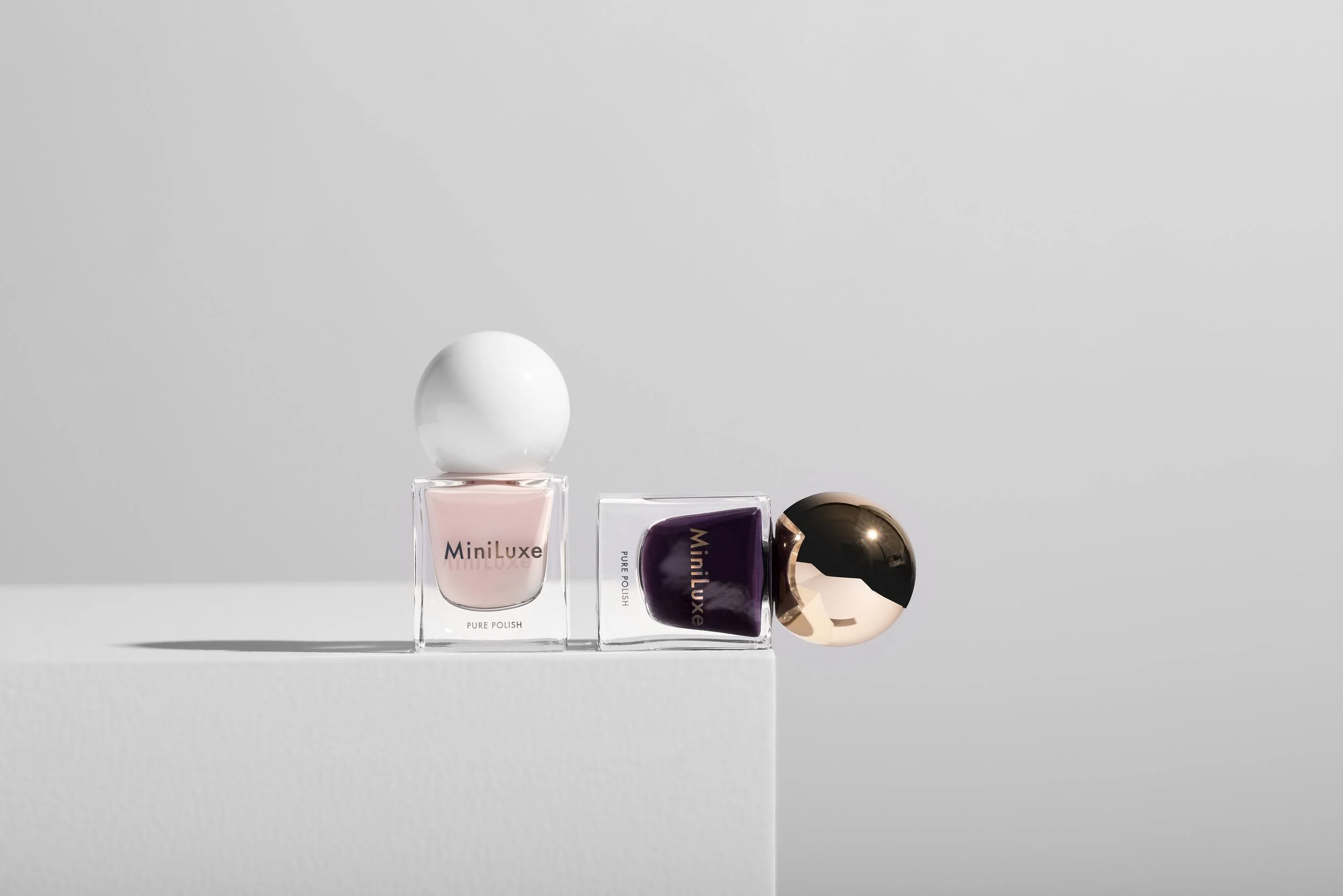 Three bottles of MiniLuxe nail polish arranged in a row on a white surface with a light gray background, featuring pink, purple, and gold caps.