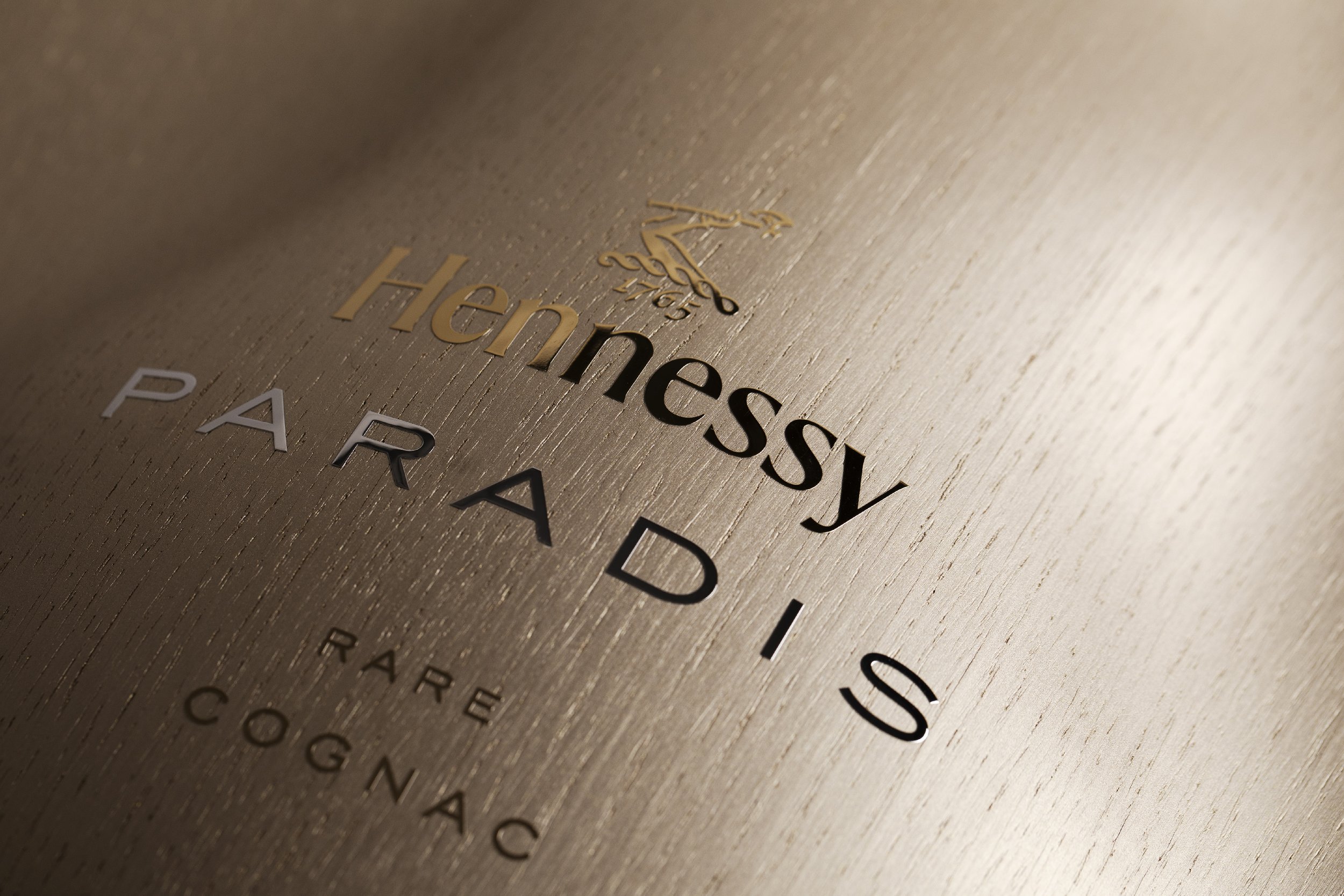 Close-up of a gold-colored surface with black and gold text and an elegant clock graphic, displaying the name 'Hennessy' and words like 'PARADIS,' indicating a luxury cognac edition.