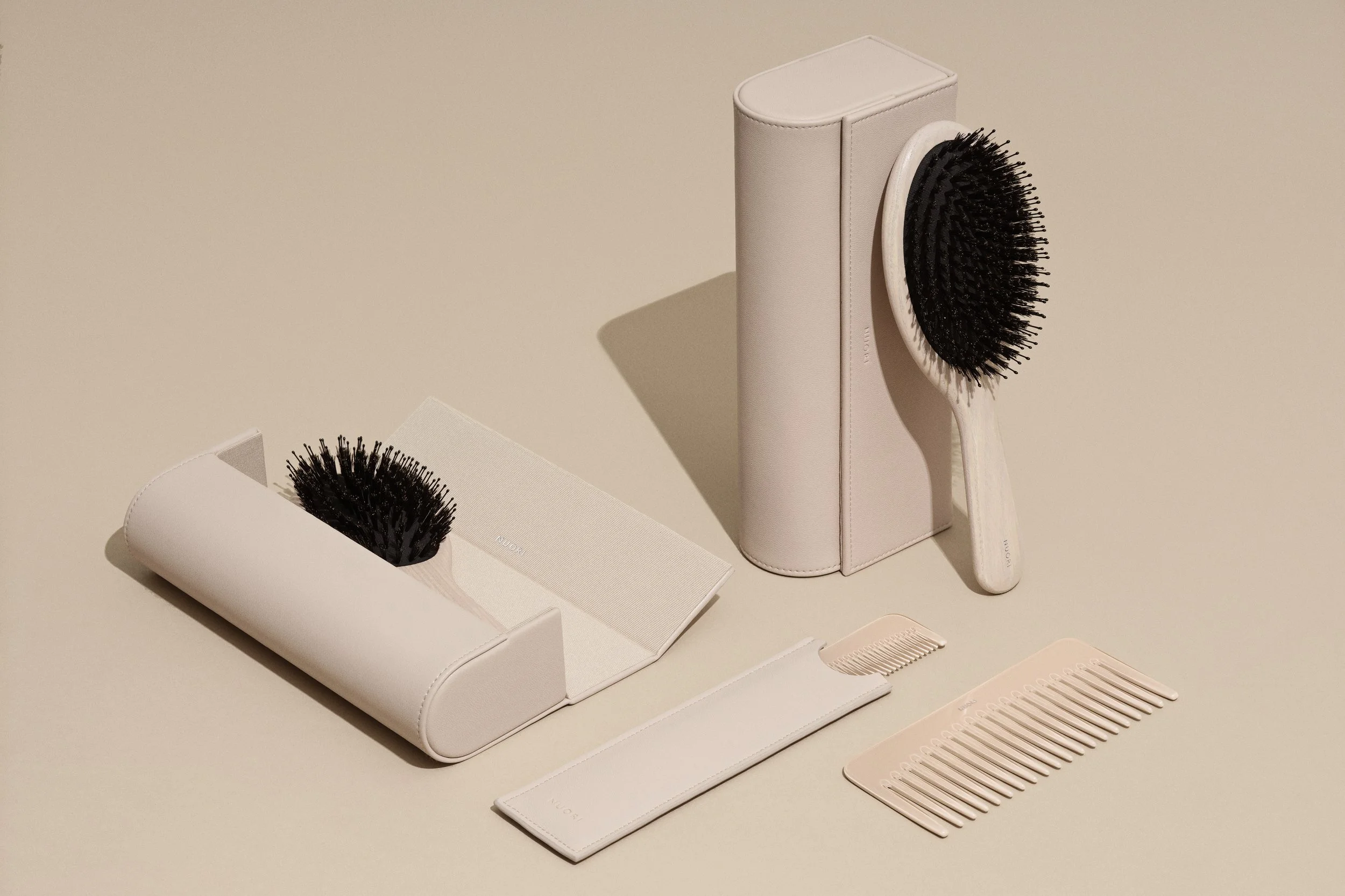 A set of hairbrushes with black bristles, including a round brush, stored in white cases and pouches, with a beige comb, all placed on a light beige surface.