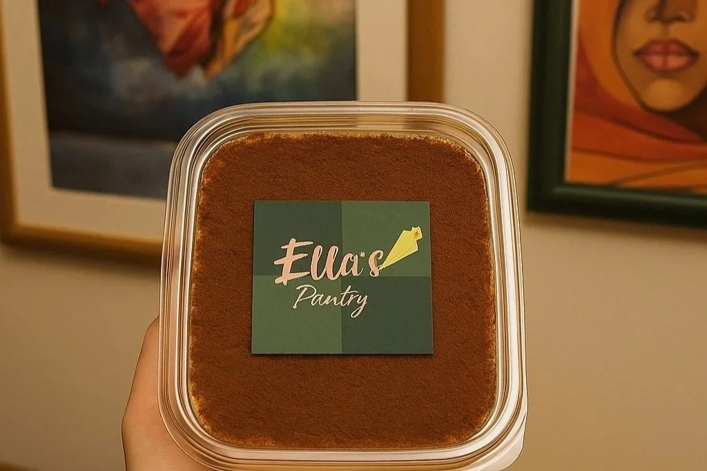 A container of tiramisu  dusted with cocoa powder with a label that reads 'Ella's Pantry', held in front of framed artwork on a wall.